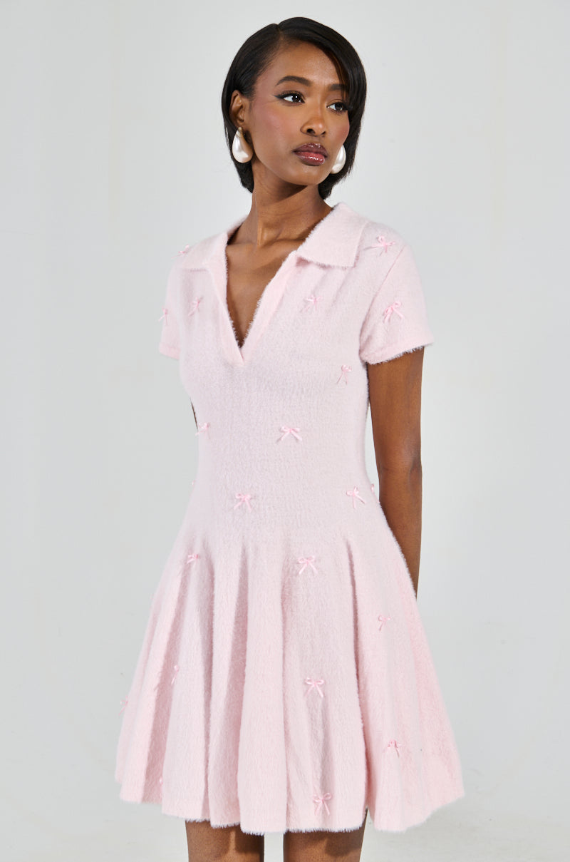 A woman wears the BOW DOWN GIRL MINI DRESS IN PINK, a light pink mini with a v-neck and bow details. The skater silhouette with a flared skirt complements her shoulder-length dark hair as she gazes to the side, set against a plain gray background.