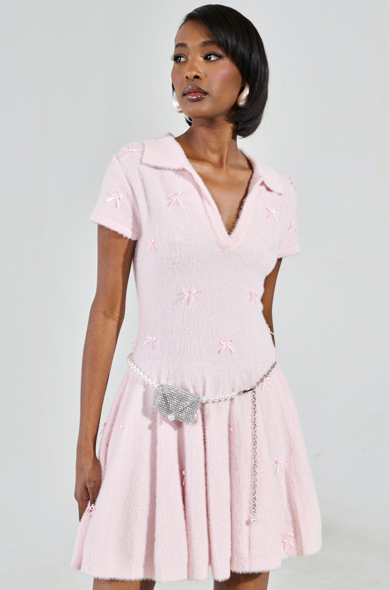 The BOW DOWN GIRL MINI DRESS IN PINK, with a mohair-like knit and small bow details, features a skater silhouette with short sleeves, a V-neck, and a flared skirt. A sparkly chain belt and earrings add flair against the neutral background, exuding confidence.