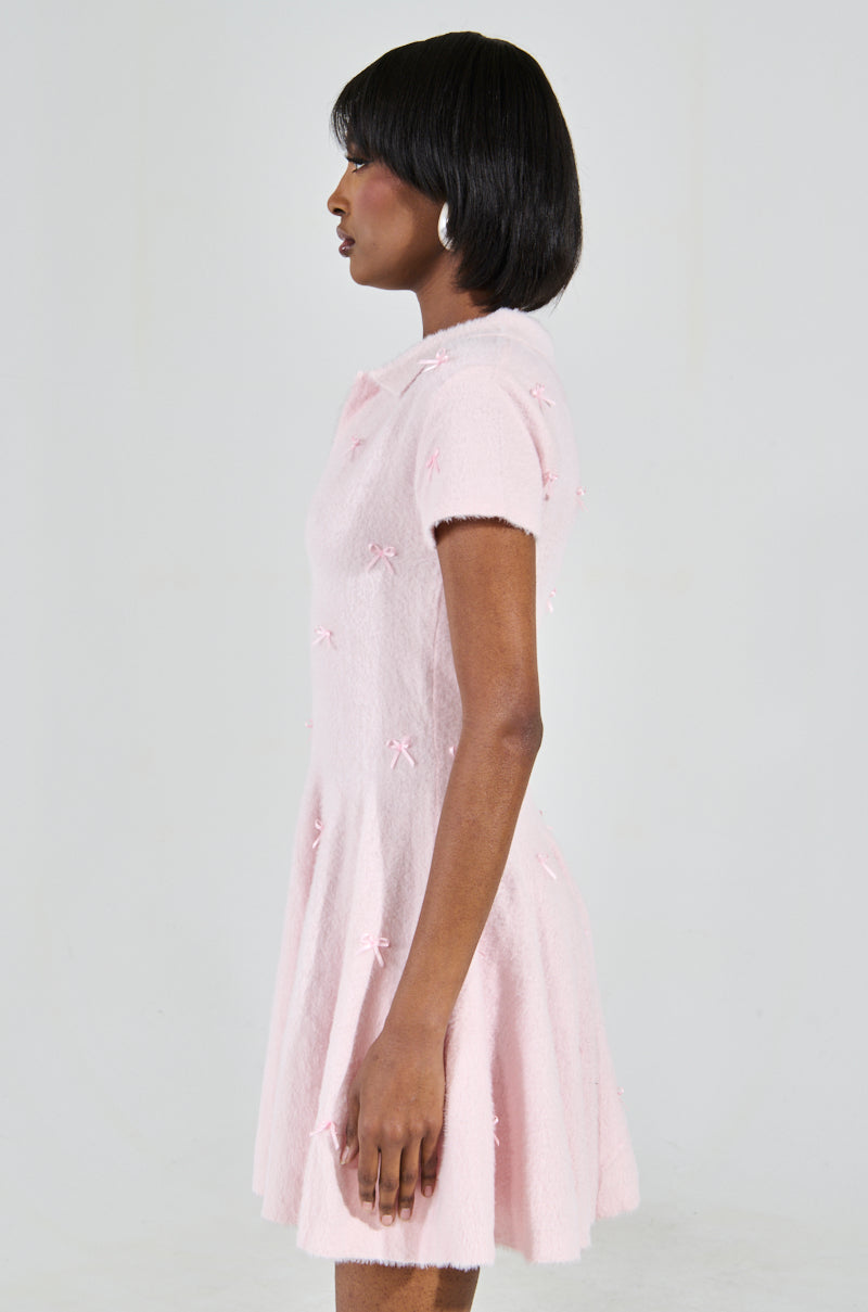 A person with short black hair is in profile wearing the BOW DOWN GIRL MINI DRESS IN PINK adorned with small bows and white hoop earrings, featuring a skater dress silhouette that adds charm against a plain light grey background.