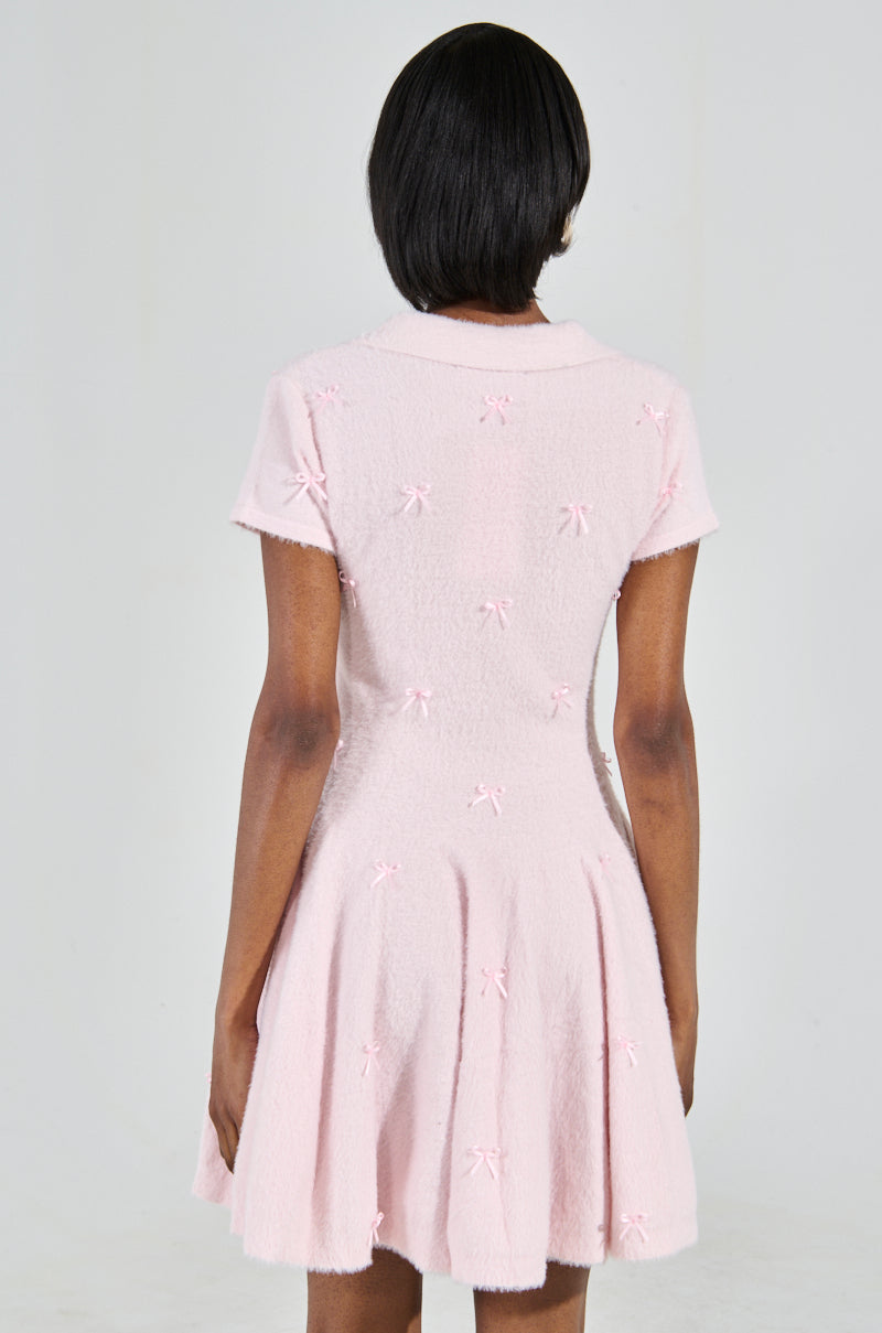 A person with short dark hair is wearing the Bow Down Girl Mini Dress in pink, featuring small bows, short sleeves, and a skater dress silhouette. They stand with their back to the camera against a plain light background.