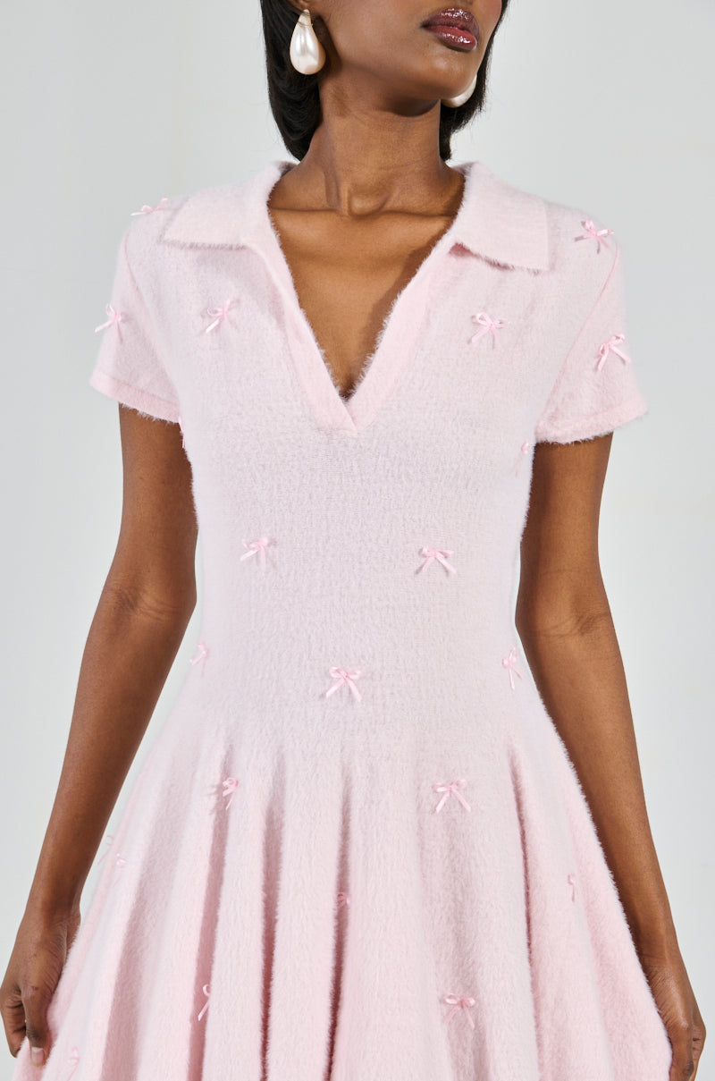 A woman elegantly showcases the Bow Down Girl Mini Dress in pastel pink, featuring a fuzzy mohair-like knit, short sleeves, a skater dress silhouette with a deep V-neck and collar, and small bow details. She complements it with pearl earrings and straight-styled hair.