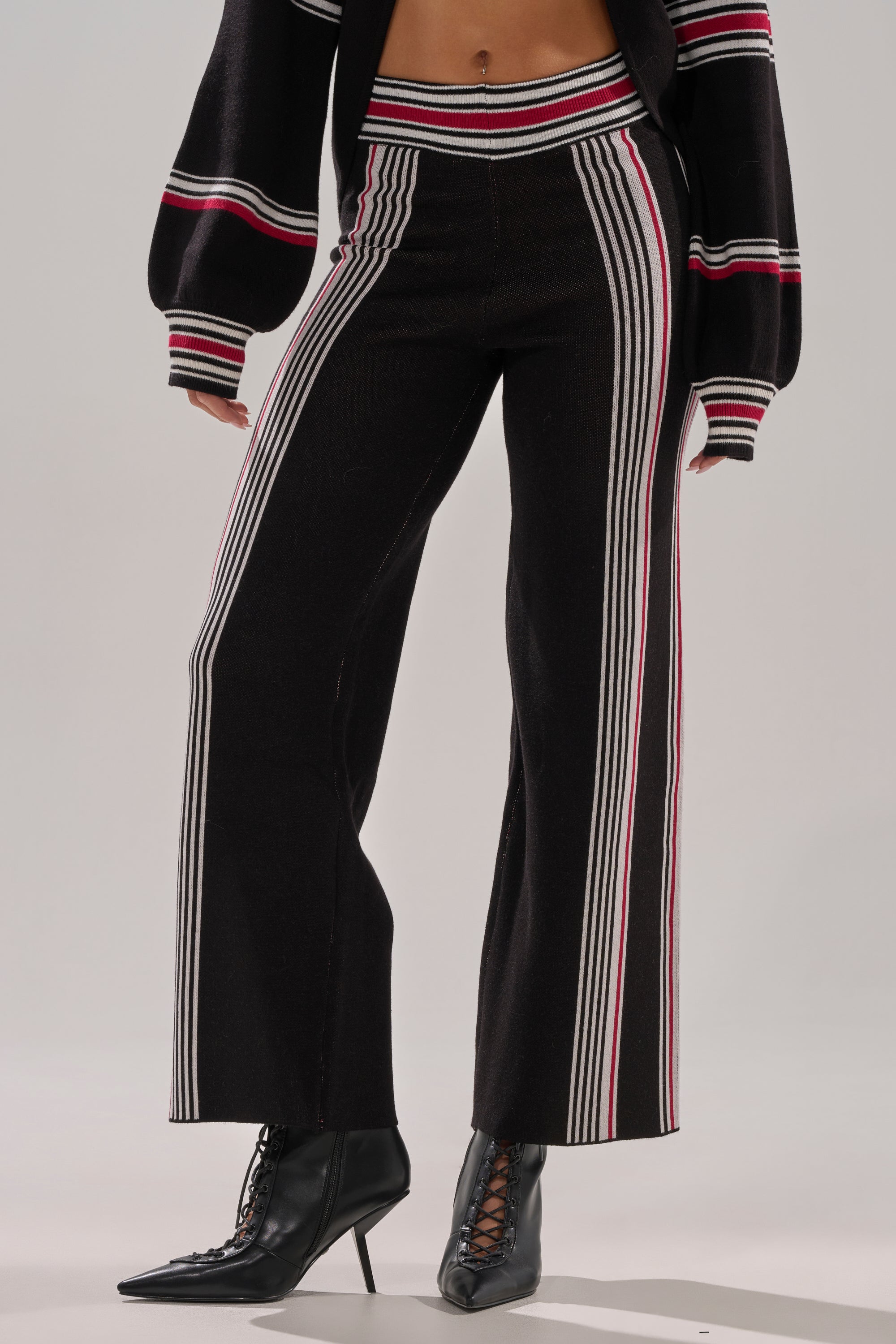 Wearing the KENDALL SWEATER WIDE LEG PANT—black high-rise, wide-leg pants with white and red stripes and a matching knit sweater with striped cuffs and hem—styled with black lace-up heeled boots against a plain light background.