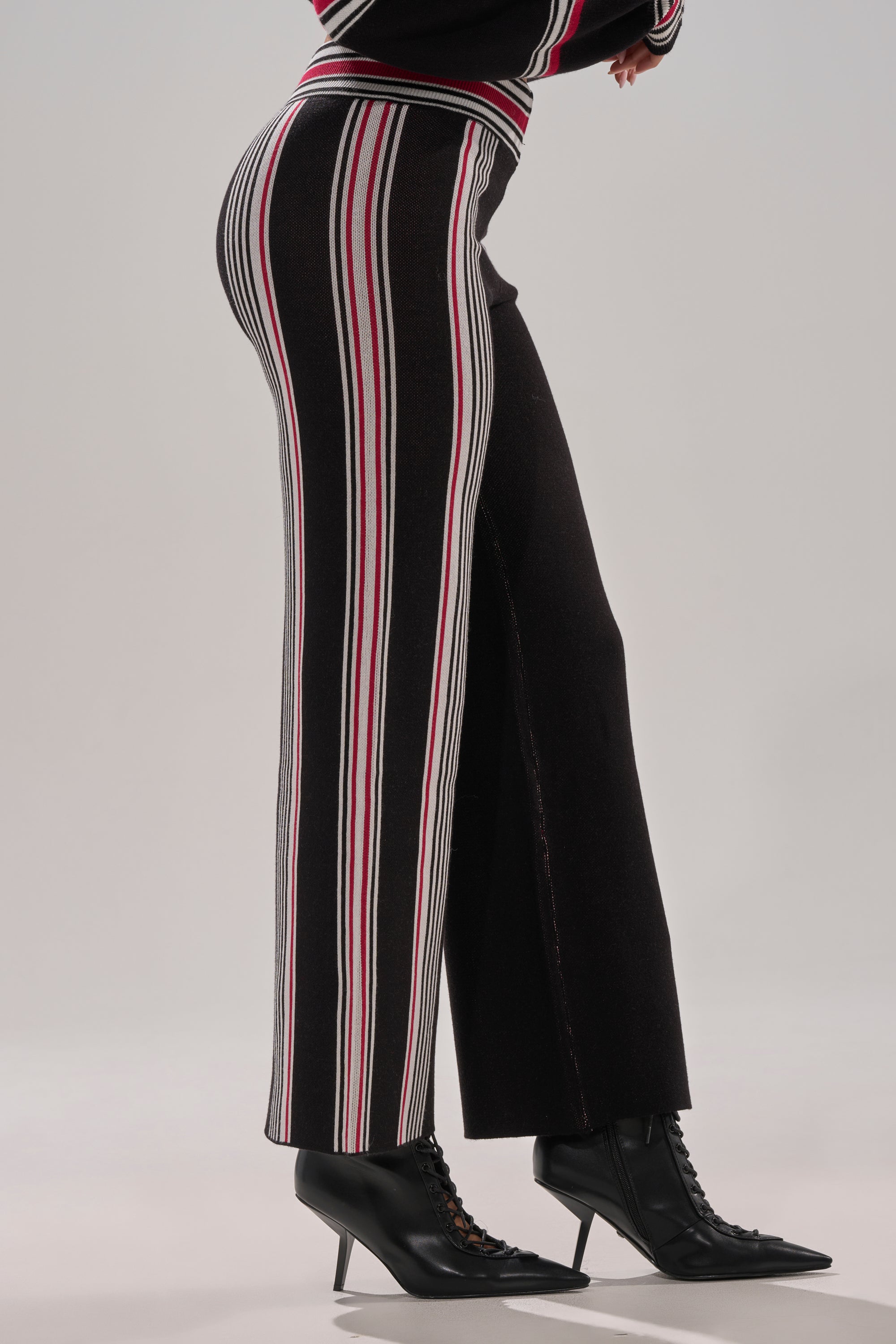 A person wearing the KENDALL SWEATER WIDE LEG PANT in high-rise black with red, white, and gray vertical stripes, paired with black pointed-toe lace-up ankle boots with high heels. The background is plain and light-colored.