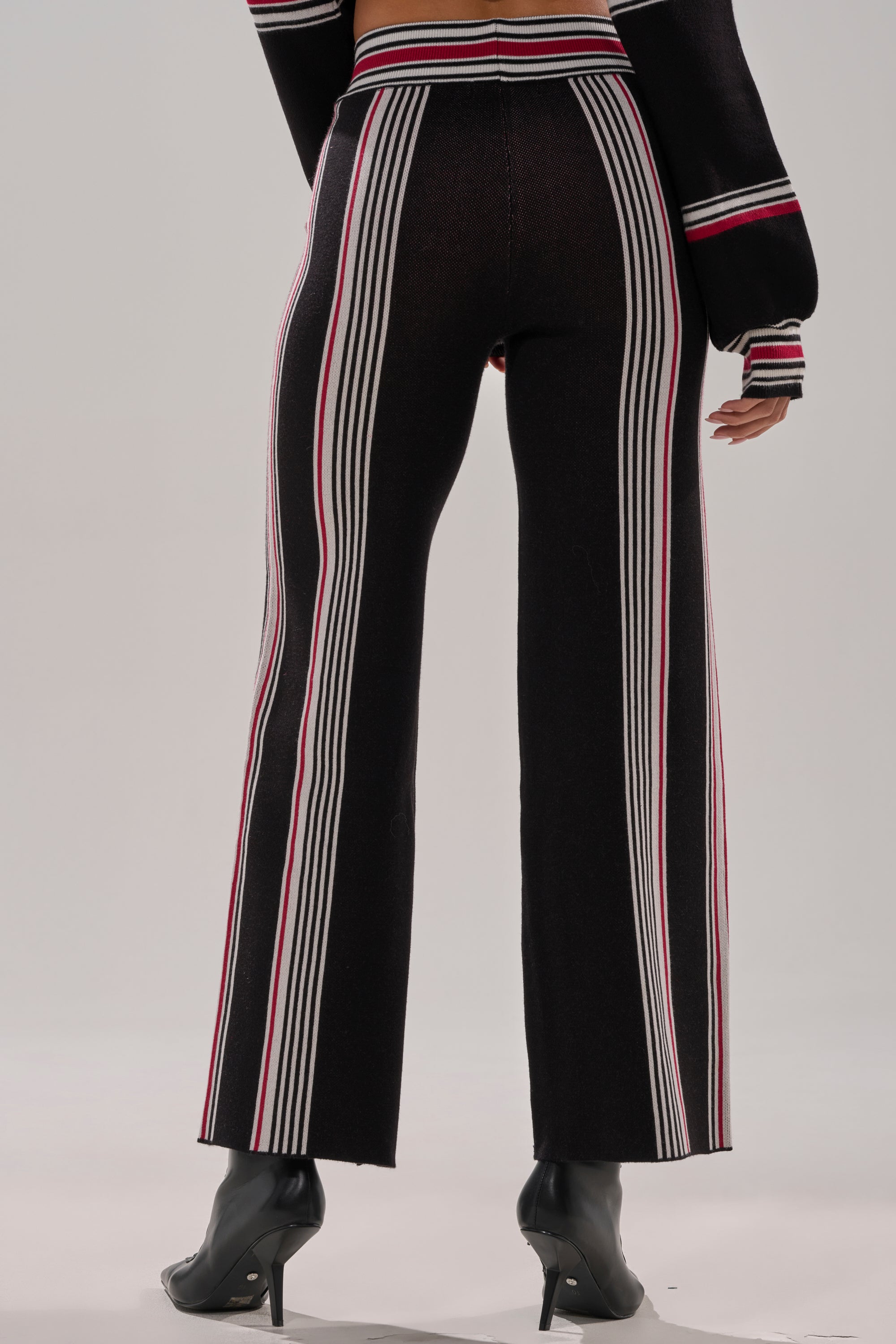 A person wears the KENDALL SWEATER WIDE LEG PANT, featuring high-rise black wide-leg pants with vertical white and red stripes and a matching knit sweater, paired with black heeled open-toe shoes against a light gray background.