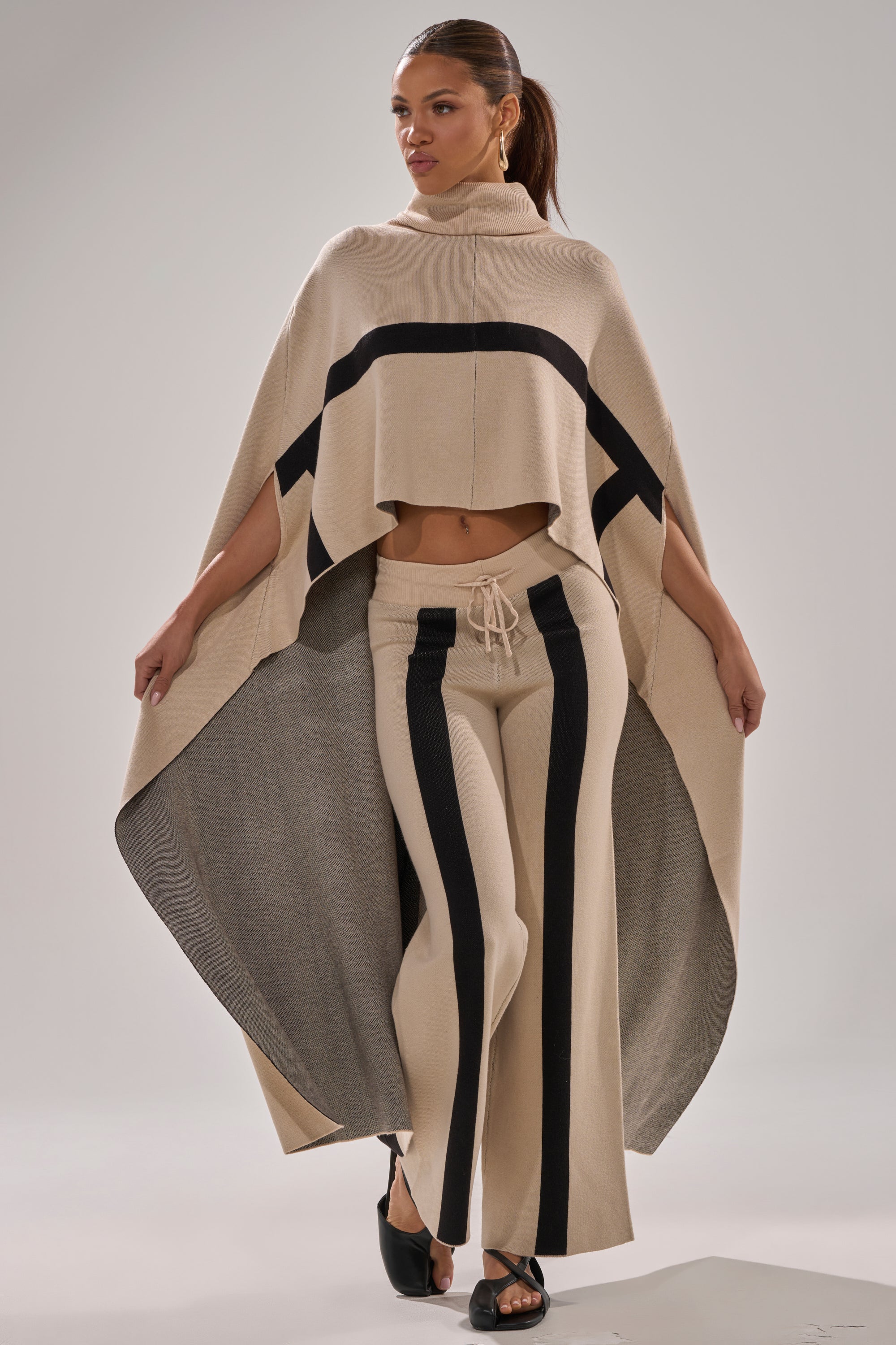 A woman models the CLAUDIA TURTLENECK SWEATER with black horizontal stripes over matching flared pants with vertical stripes and black open-toe heels, posing with arms outstretched against a plain background.