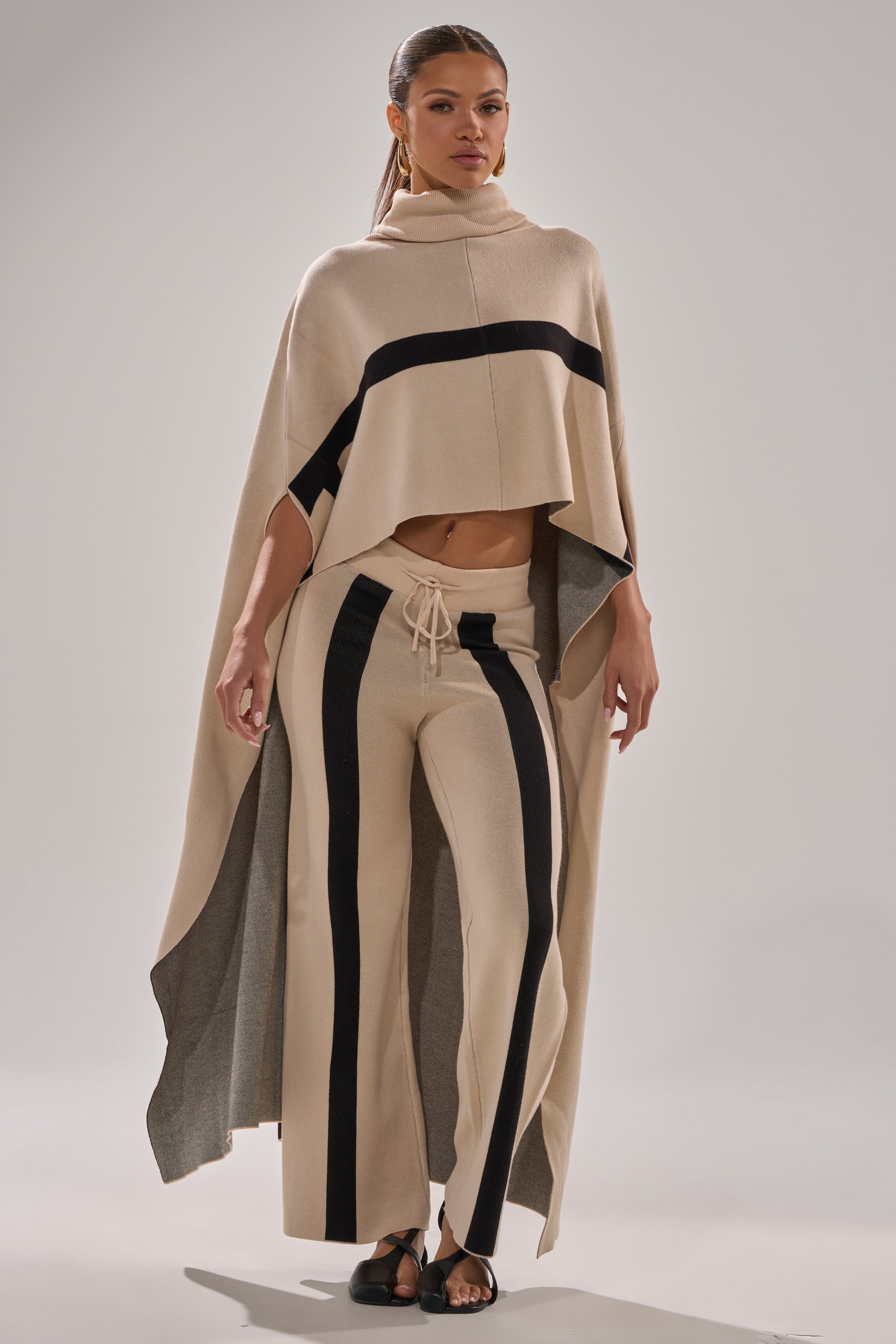 A woman poses in a beige CLAUDIA TURTLENECK SWEATER with black stripes, paired with wide-leg drawstring pants and black sandals, her hair pulled back, standing against a plain background.