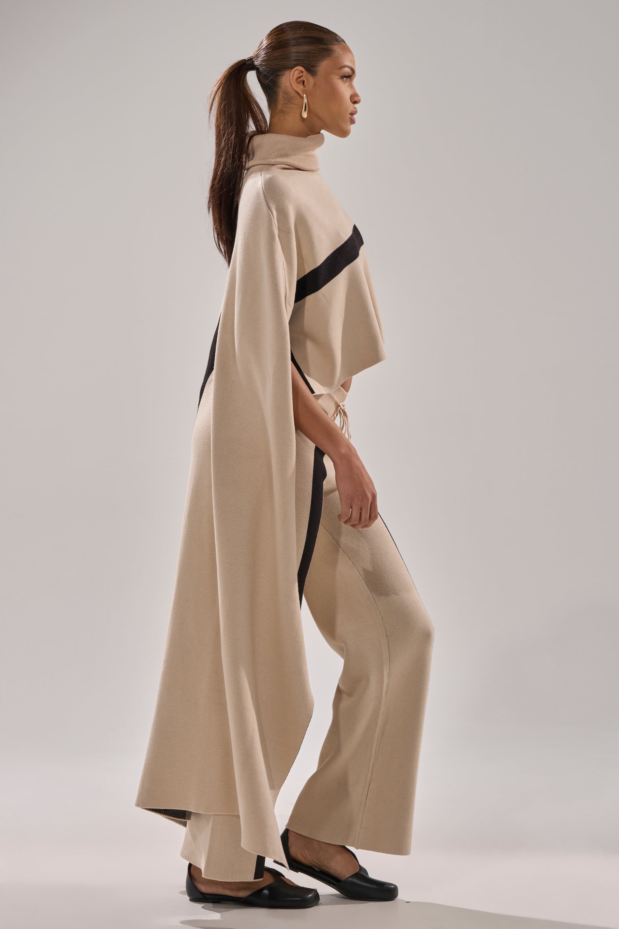 A woman in profile wears a beige CLAUDIA TURTLENECK SWEATER with black accents, wide-leg pants, and black shoes. Her hair is in a ponytail, and she sports statement earrings against a plain light background.