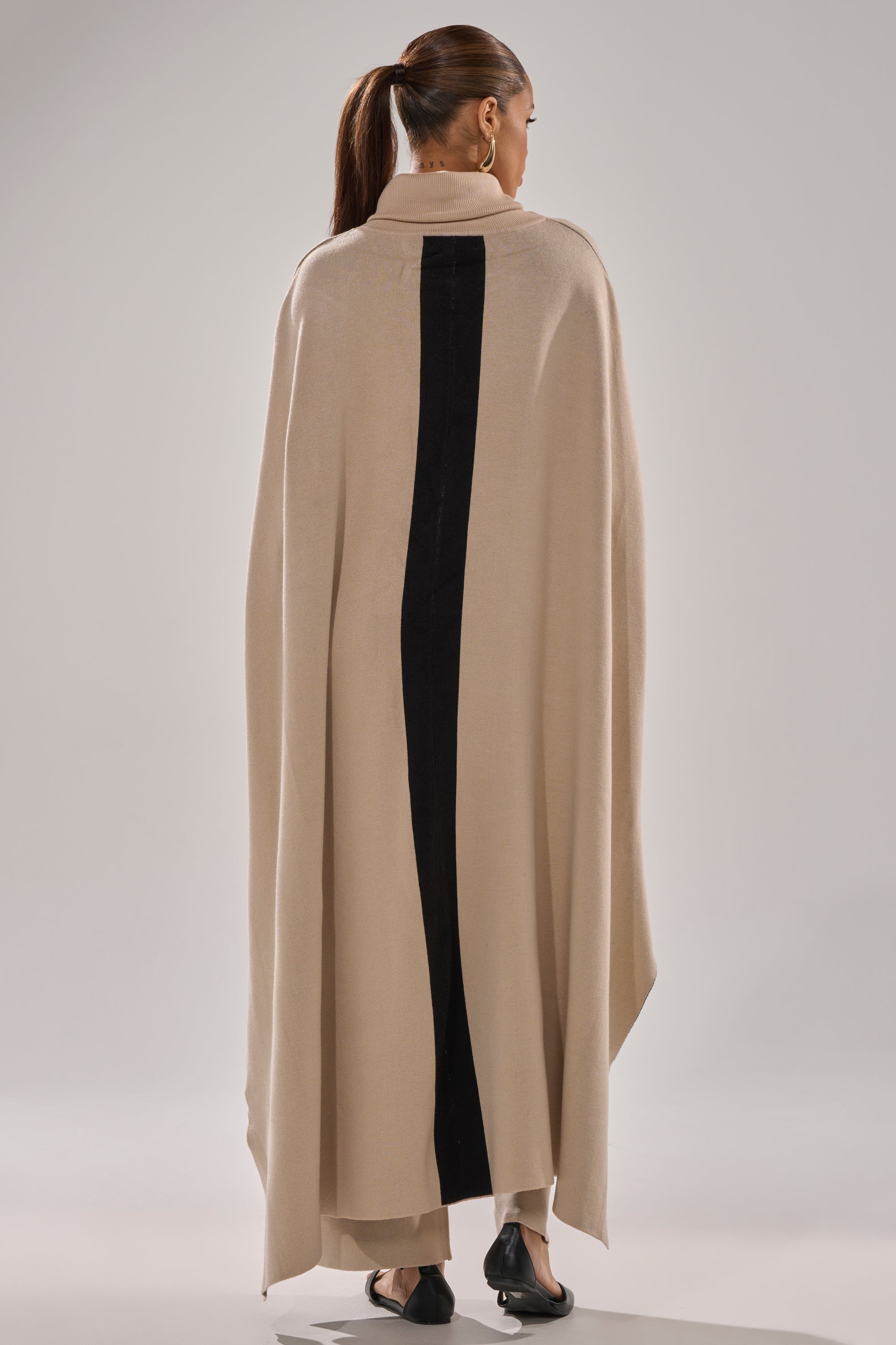 A person stands facing away, wearing the CLAUDIA TURTLENECK SWEATER with a bold black vertical center stripe, tan pants, black shoes, and a ponytail. The background is neutral and plain.