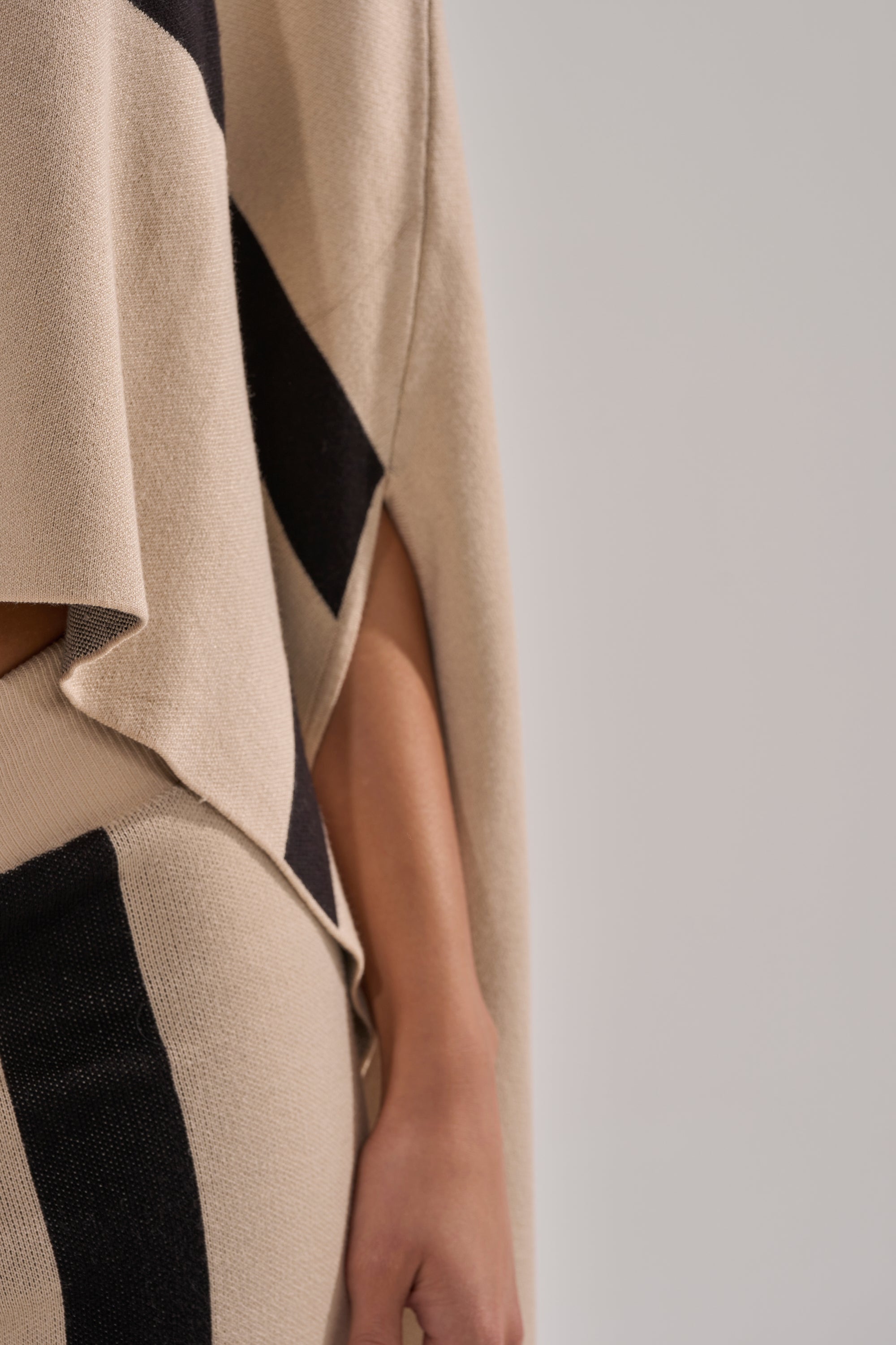 A close-up of a person wearing the CLAUDIA TURTLENECK SWEATER in beige and black, featuring draped fabric on the arm, hand, and part of the torso. The background is plain and light-colored.