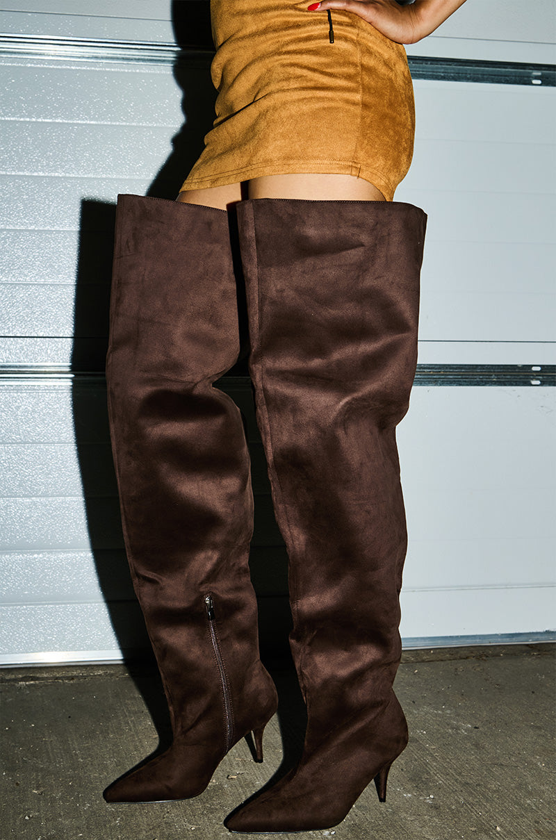 A person, shown from the waist down, stands against a garage door wearing an above-the-knee tan suede skirt and AZALEA WANG HALINA CHOCOLATE THIGH HIGH BOOT with a stiletto heel.