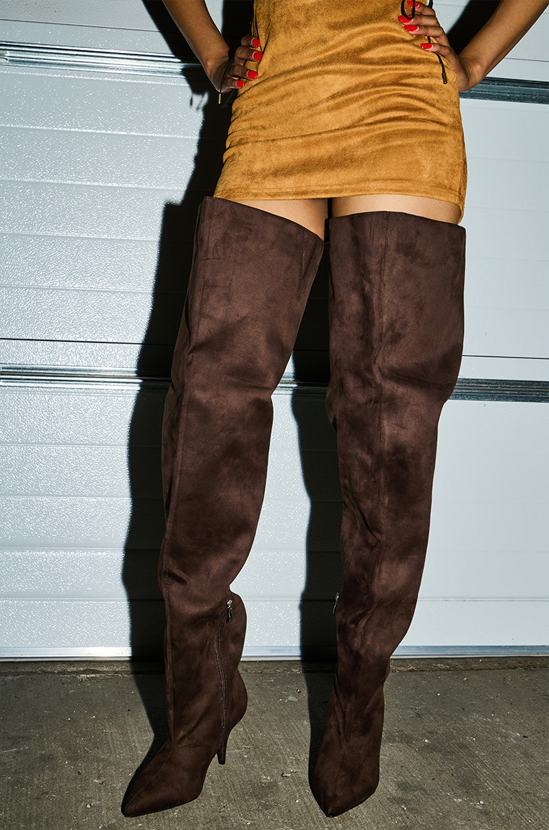 A person stands in front of a metallic garage door, hands on hips with red nails showing, wearing a tan faux suede mini dress and AZALEA WANG HALINA CHOCOLATE THIGH HIGH BOOTS.
