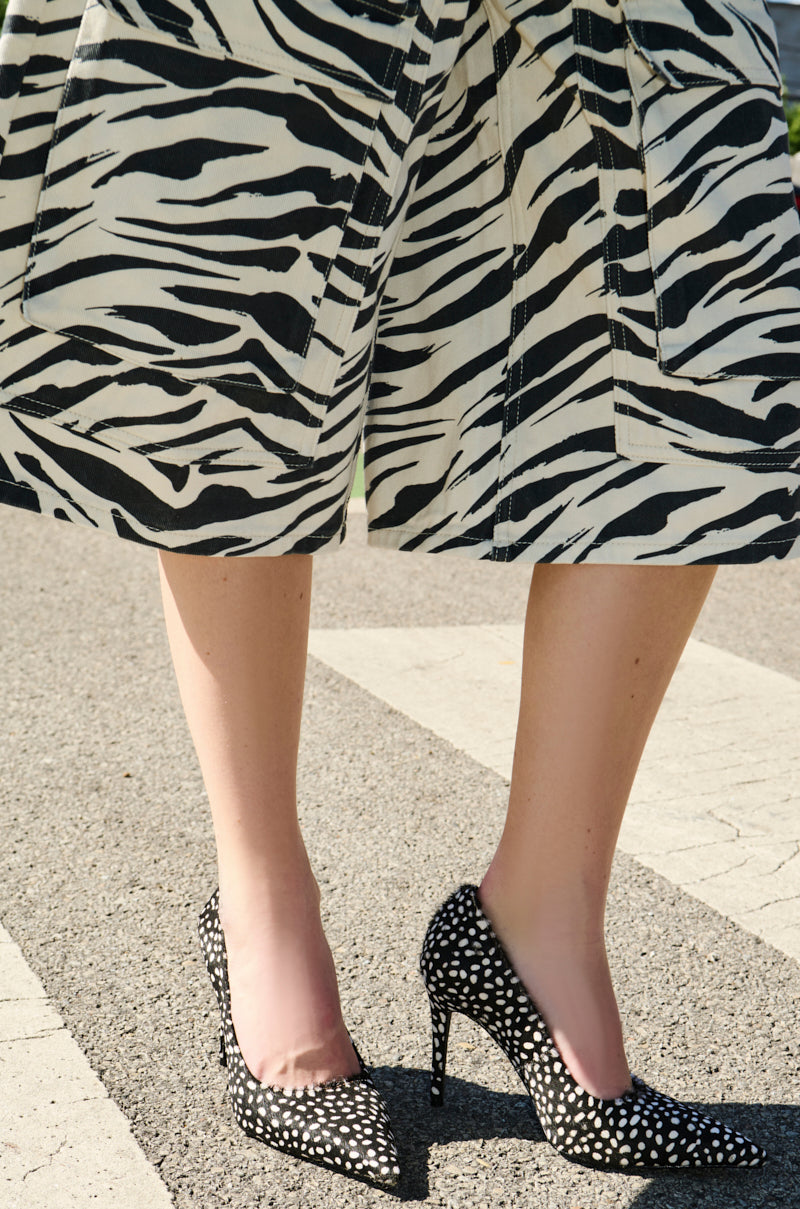 A person stands outdoors on a paved surface, showing only their legs while wearing a zebra-print skirt and the AZALEA WANG HALL BLACK MULTI SPOTTED PONY HAIR PUMP with stiletto heels and pointed toes.