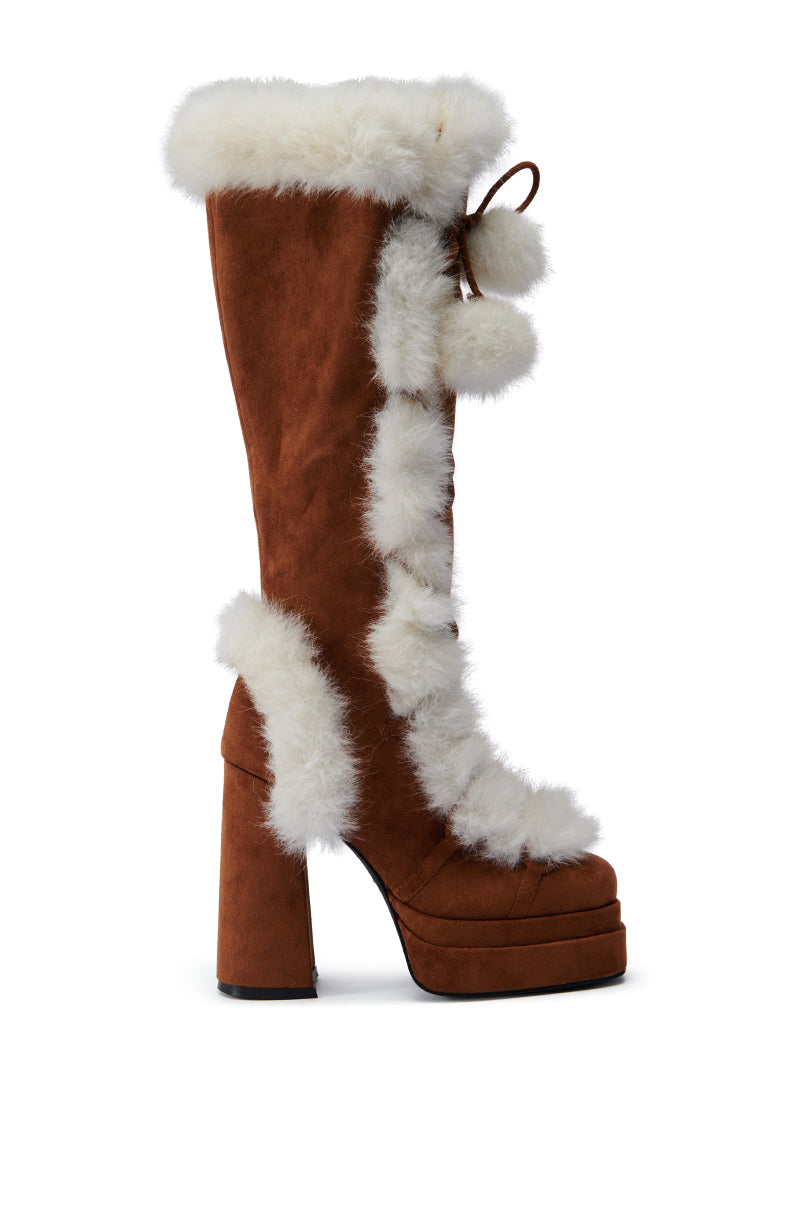 A side view of the AZALEA WANG HARIBO TAN FUR BOOT reveals its brown faux suede knee-high design, featuring a chunky platform heel. The boot is adorned with white faux fur trim running along the front, top, and back, complemented by playful pom-pom lace detailing. The backdrop is a plain white setting.