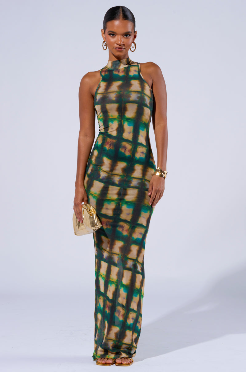 A woman stands confidently in the COOL FOR THE SUMMER PRINTED MAXI DRESS, a sleeveless, high-neck, floor-length piece adorned with a green, brown, and yellow tie-dye pattern. She accessorizes with a gold clutch in her right hand, complemented by gold hoop earrings and bracelets. Her confident expression shines against the plain background.