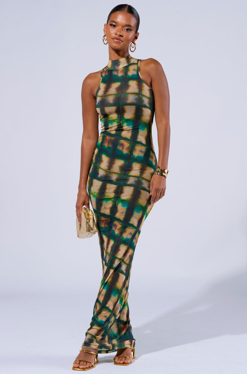 A person stands confidently in a sleeveless, floor-length COOL FOR THE SUMMER PRINTED MAXI DRESS adorned with a green and gold tie-dye pattern. They accessorize with a small gold clutch, gold hoop earrings, and a gold bracelet. Their hair is elegantly pulled back, and they complete the look with strappy heels.