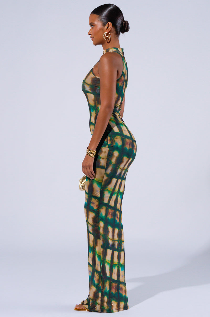 A woman stands in profile against a light gray background, wearing the form-fitting, sleeveless COOL FOR THE SUMMER PRINTED MAXI DRESS in shades of green, brown, and blue. She holds a small gold clutch and wears gold hoop earrings and bracelets. Her hair is pulled back in a sleek bun.