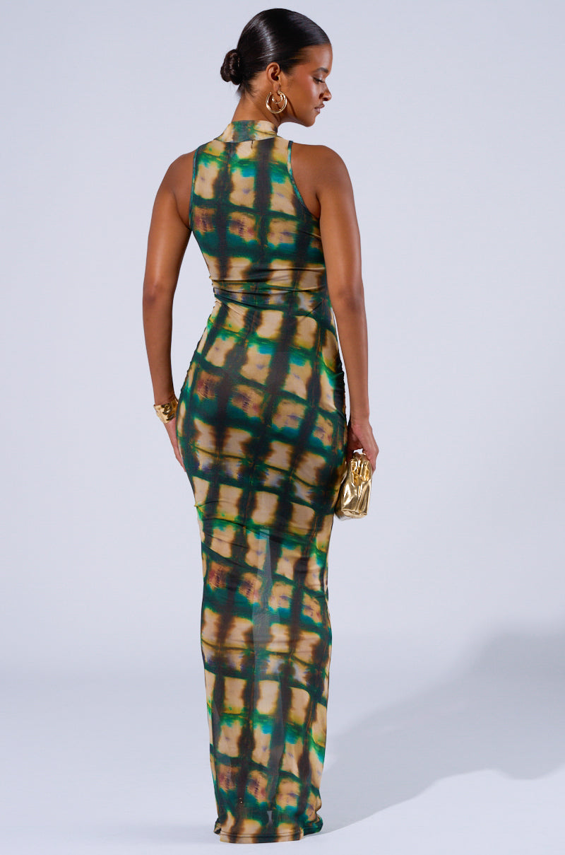 A woman is seen from the back wearing the COOL FOR THE SUMMER PRINTED MAXI DRESS, featuring a bodycon fit in a green, brown, and yellow tie-dye pattern. Her hair is styled in a sleek bun. She holds a gold clutch bag in her left hand and wears matching accessories, standing against a plain background.
