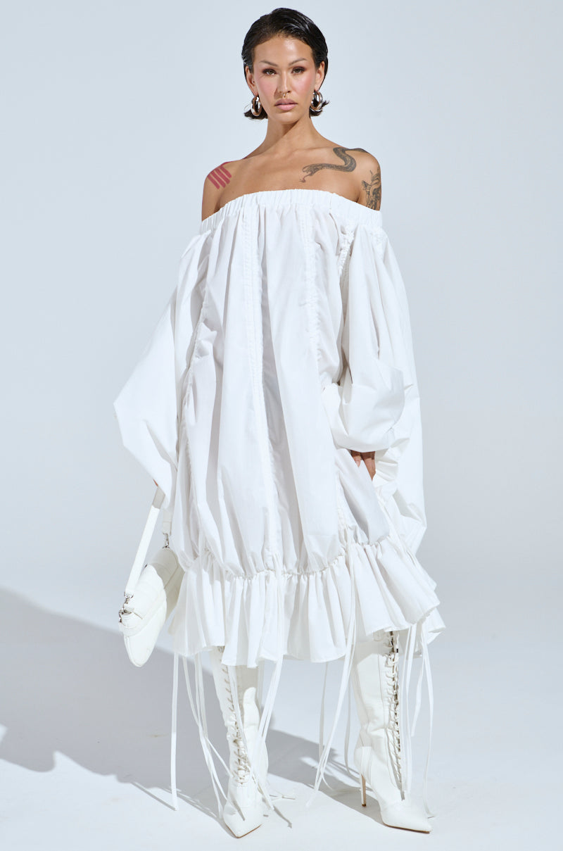 Wearing the FREE AS CAN BE MINI DRESS IN WHITE, a person stands elegantly, completing the look with knee-high white lace-up boots. Tattoos on their shoulder and earrings enhance the style against a plain, light background.