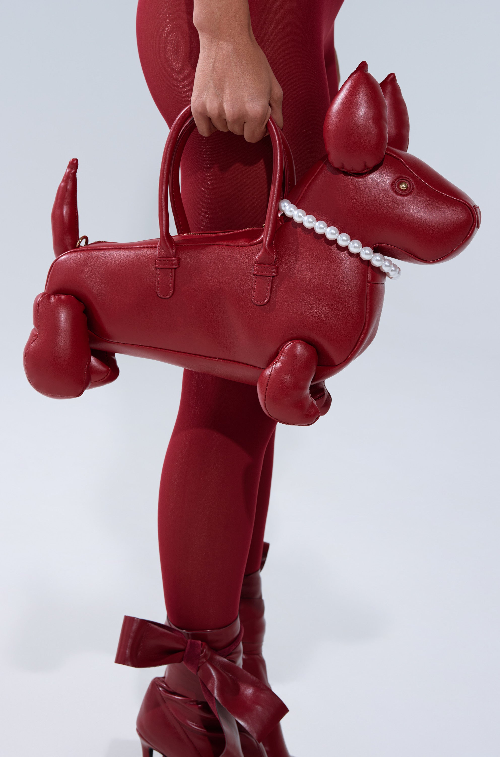 A person in red holds the PRETTY BUT I BITE BACK PURSE, a dog-shaped bag with a pearl collar, styled with red leggings and ribbon-tied heeled shoes.