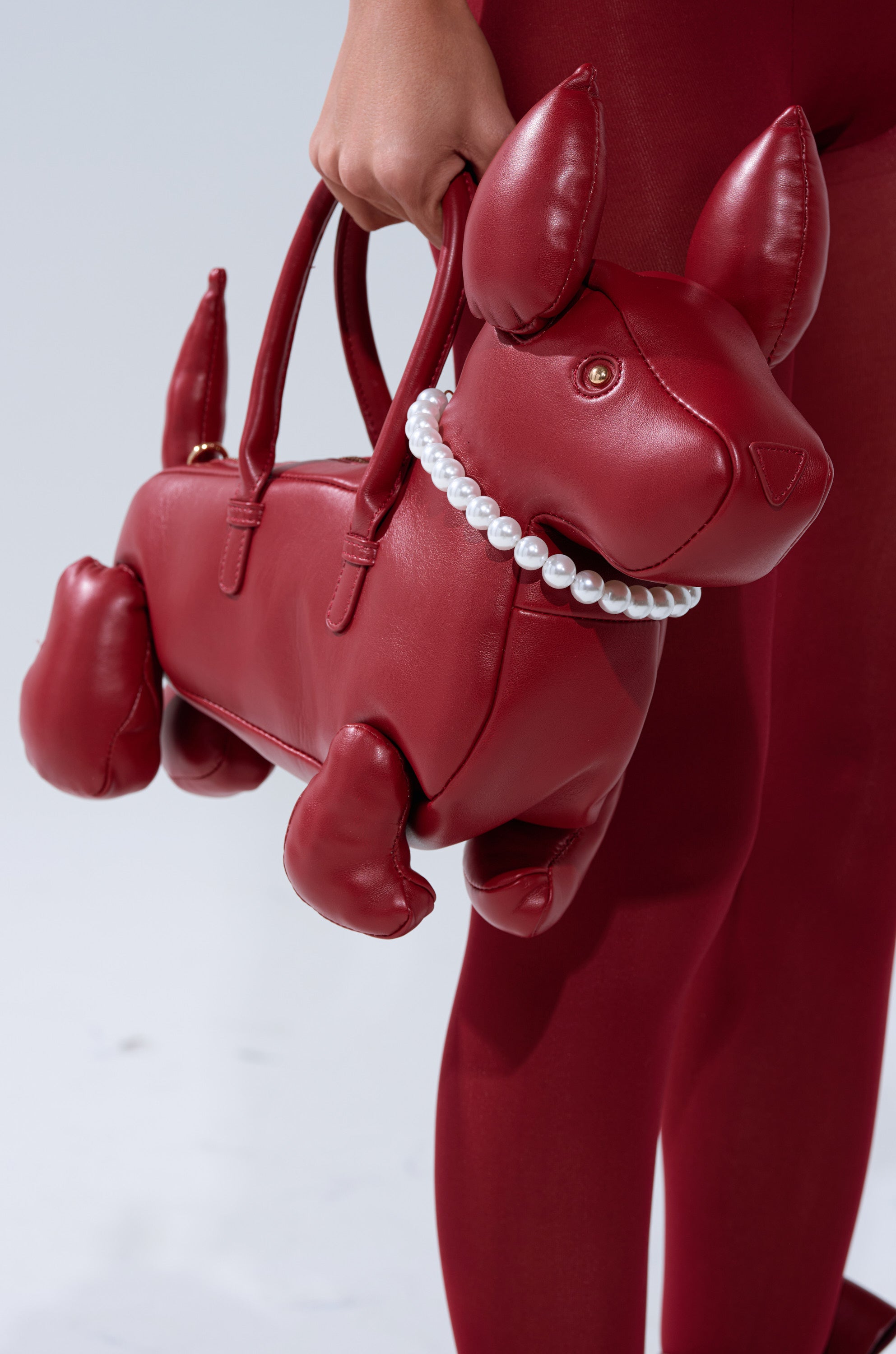 A person in red holds the PRETTY BUT I BITE BACK PURSE, a dog-shaped bag with a pearl necklace detail around its neck and a detachable shoulder strap.