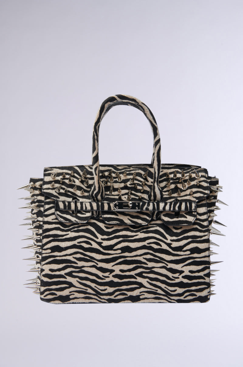 The TRY ME EMBELLISHED TOTE, a zebra print handbag with two handles and bold metallic spike accents along the edges and top, is displayed against a plain light background.