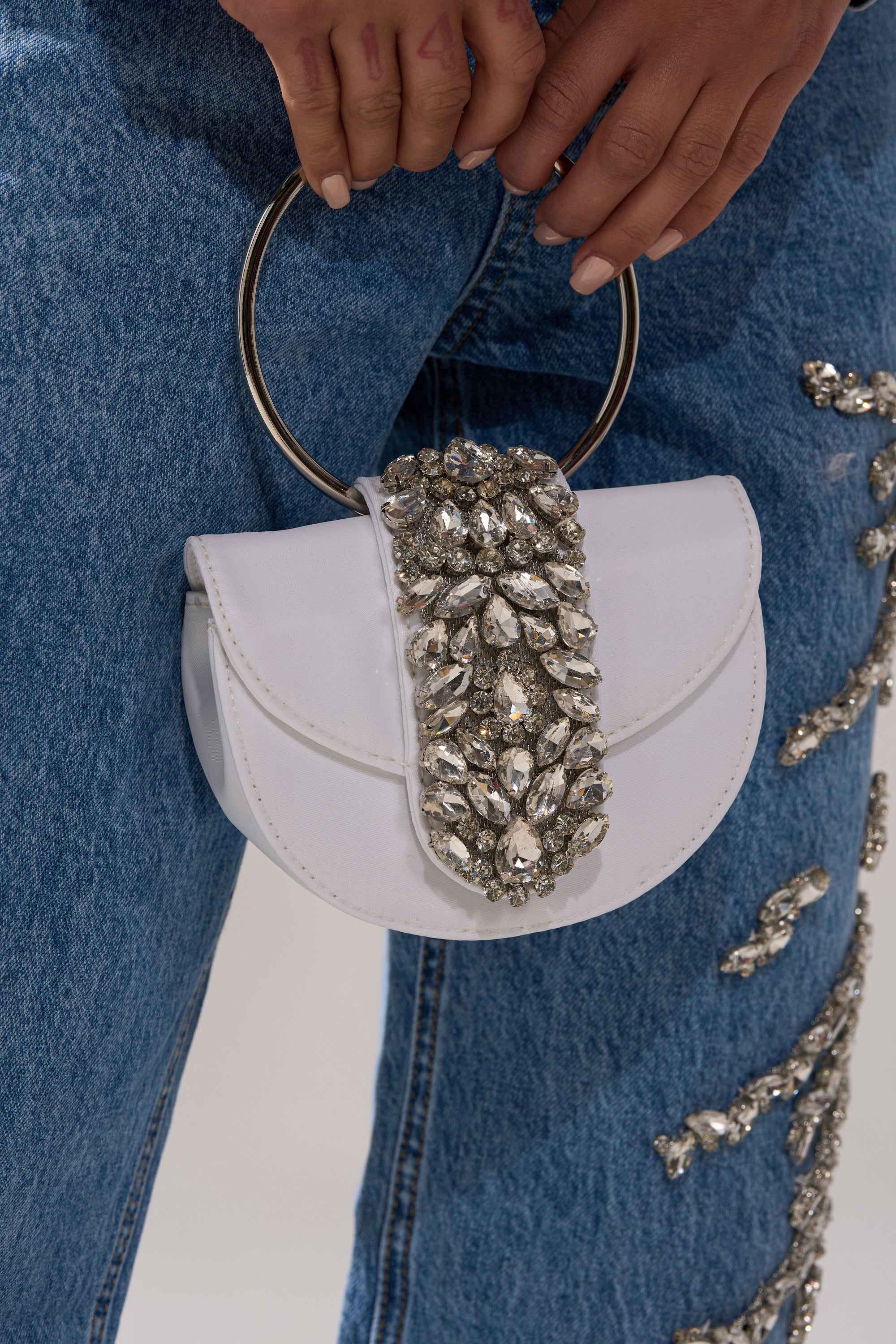 A person in blue jeans holds the TO THE EVENT EMBELLISHED PURSE—a small white handbag with silver rhinestones and a metal ring handle—making it an eye-catching accessory for any event.