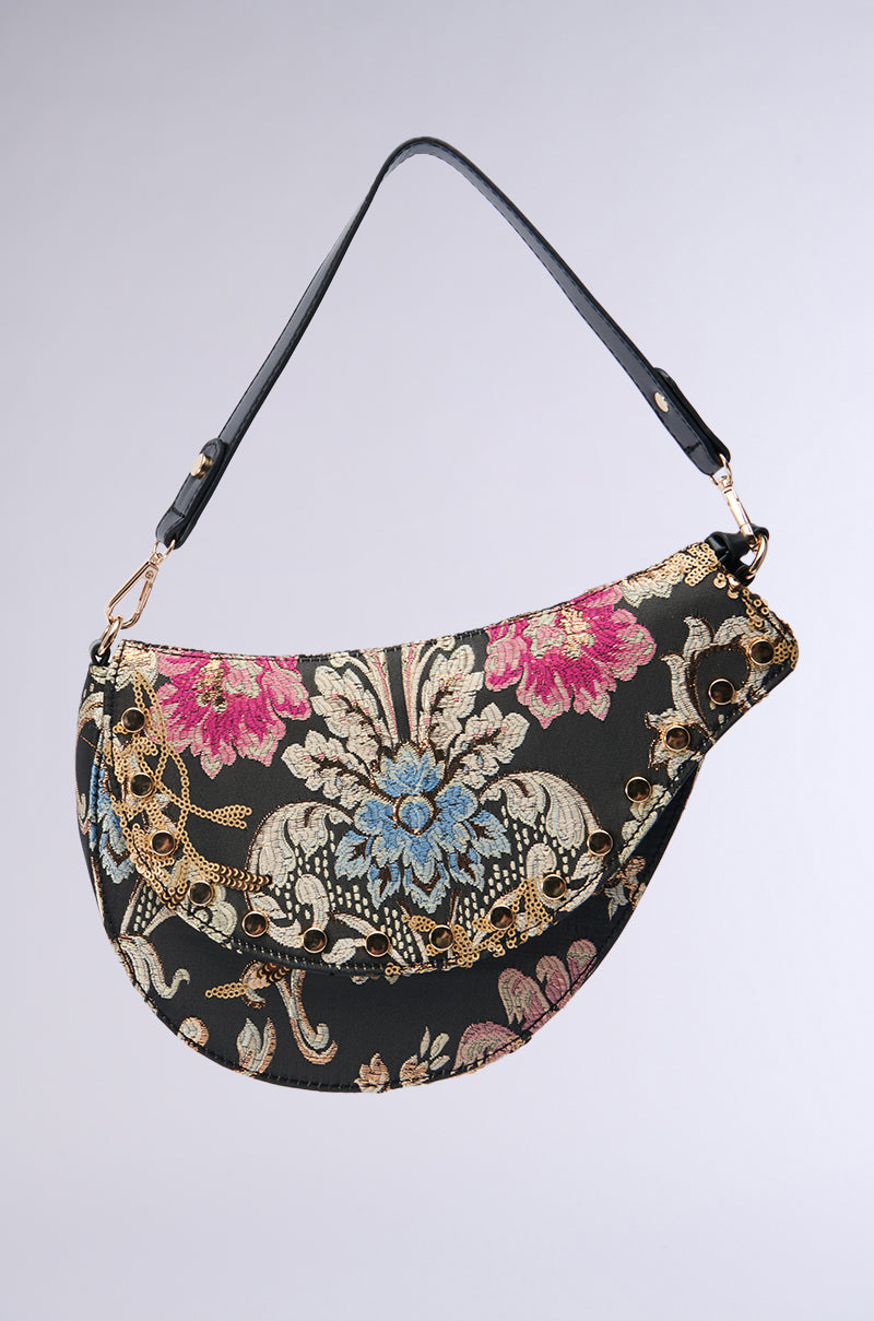 Introducing the BROCADE DREAMS SADDLE BAG, a crescent-shaped accessory featuring a black faux leather handle. Its dark background is adorned with an intricate floral pattern in pink, blue, and gold. The design is completed with modern gold eyelet details and metallic accents.