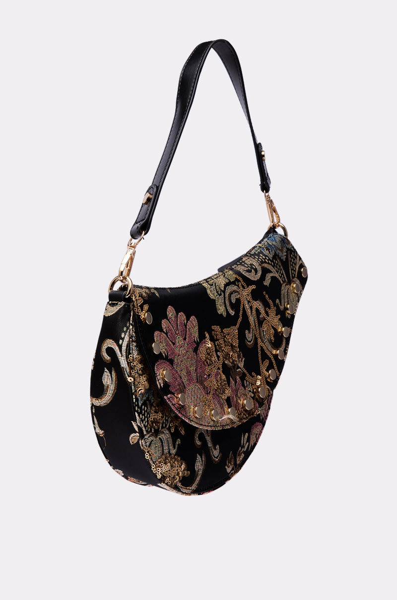 The BROCADE DREAMS SADDLE BAG is a crescent-shaped brocade accessory with a black base, featuring gold, pink, and green floral embroidery and a structured design. It has a black strap embellished with metallic accents for an elegant touch.