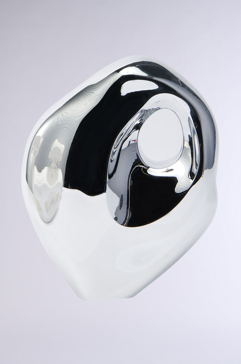 The SO MOD PURSE is a shiny, abstract silver sculpture with smooth, rounded surfaces and a central hole. Its design mimics a reflective silver handbag set against a plain gray background, creating varying shades of silver and gray.