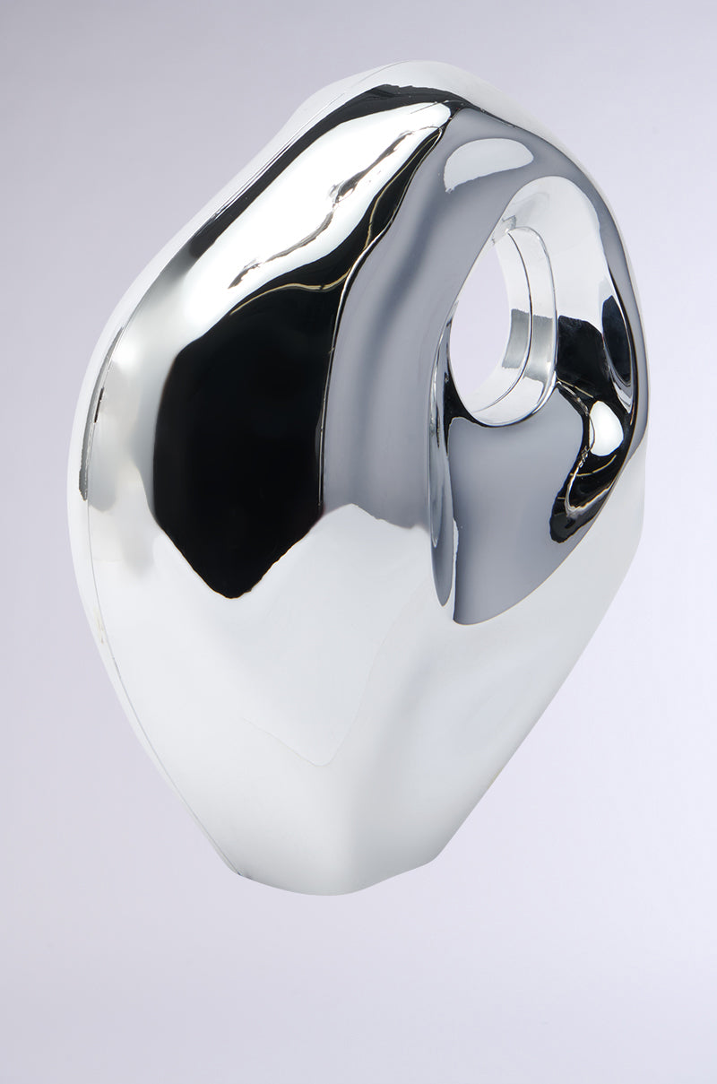 The SO MOD PURSE is a sleek, polished metallic bag with an organic shape, featuring a central circular hole. Its high-gloss surface resembles a sculpted art piece, reflecting light to create dynamic highlights and contrasts against a neutral background.