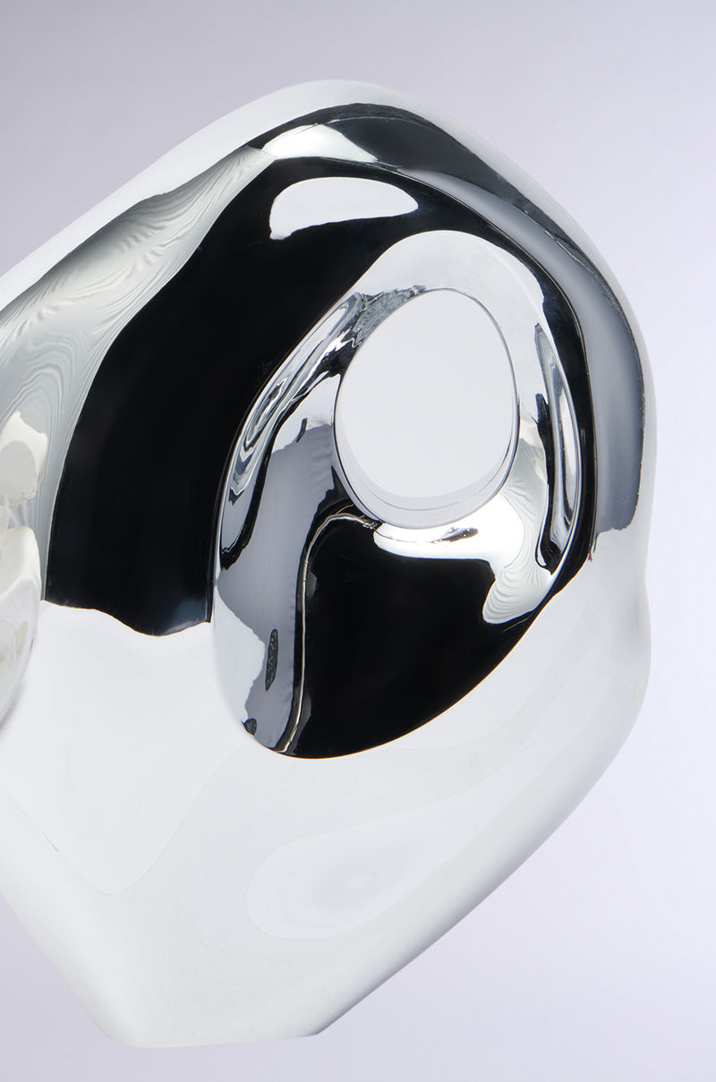 The SO MOD PURSE features a shiny metallic design, reminiscent of abstract sculpture, with smooth curves and a central oval opening. Its reflective surface distorts reflections against a neutral gray background, resembling sculpted art.