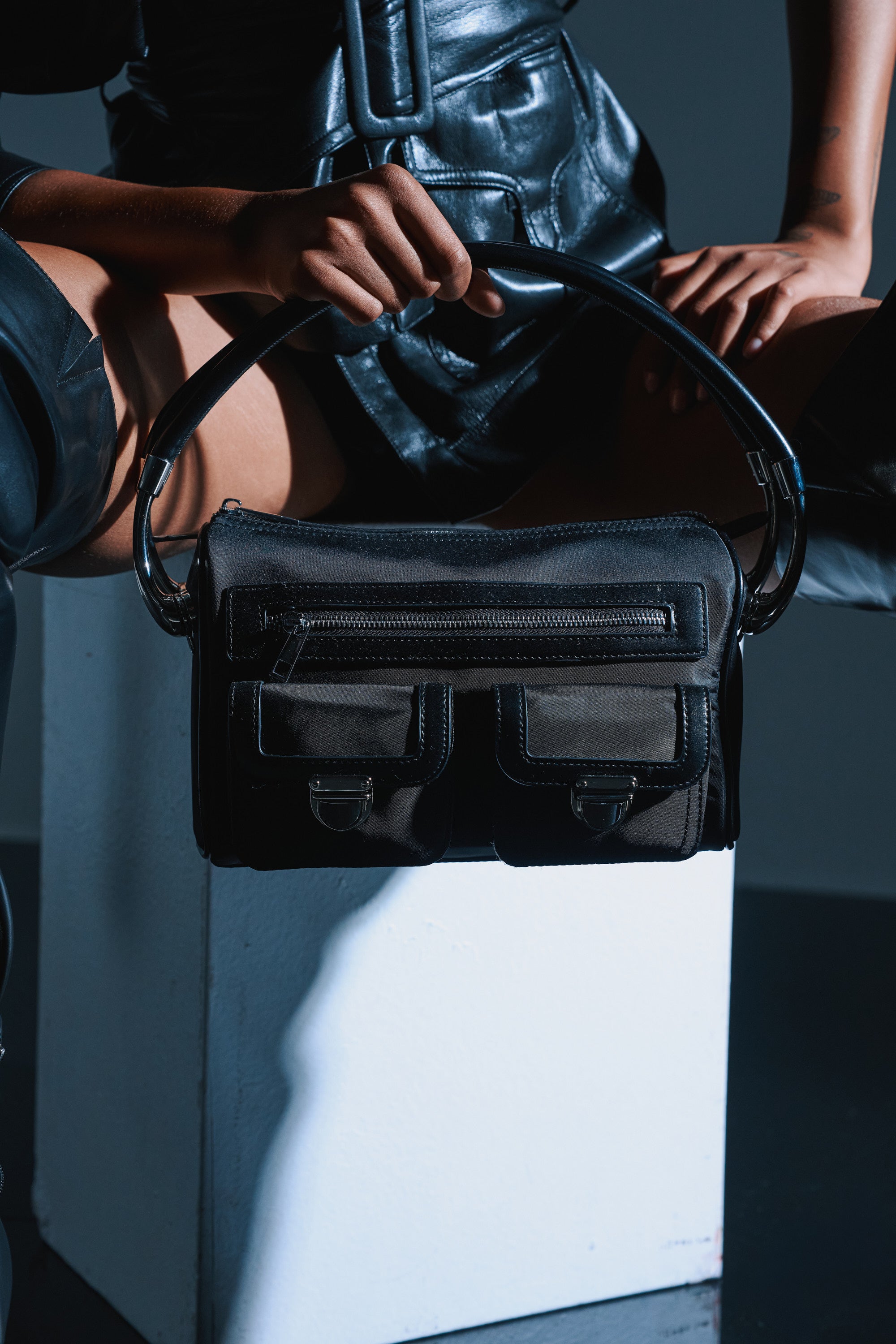 A person in a black leather outfit sits on a white block, holding the REALLY LIKE THAT PURSE with zippers and front pockets. The dark background highlights the purse and outfit, creating a striking modern look.