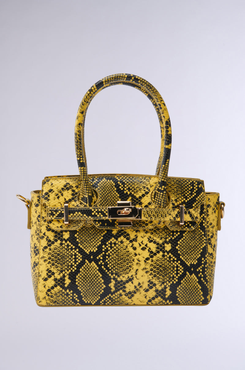 The NEVER TYPICAL SNAKE PRINT PURSE is a yellow and black handbag with a snakeskin pattern, gold-tone hardware, a top handle, and a front lock closure, displayed against a light gray background.