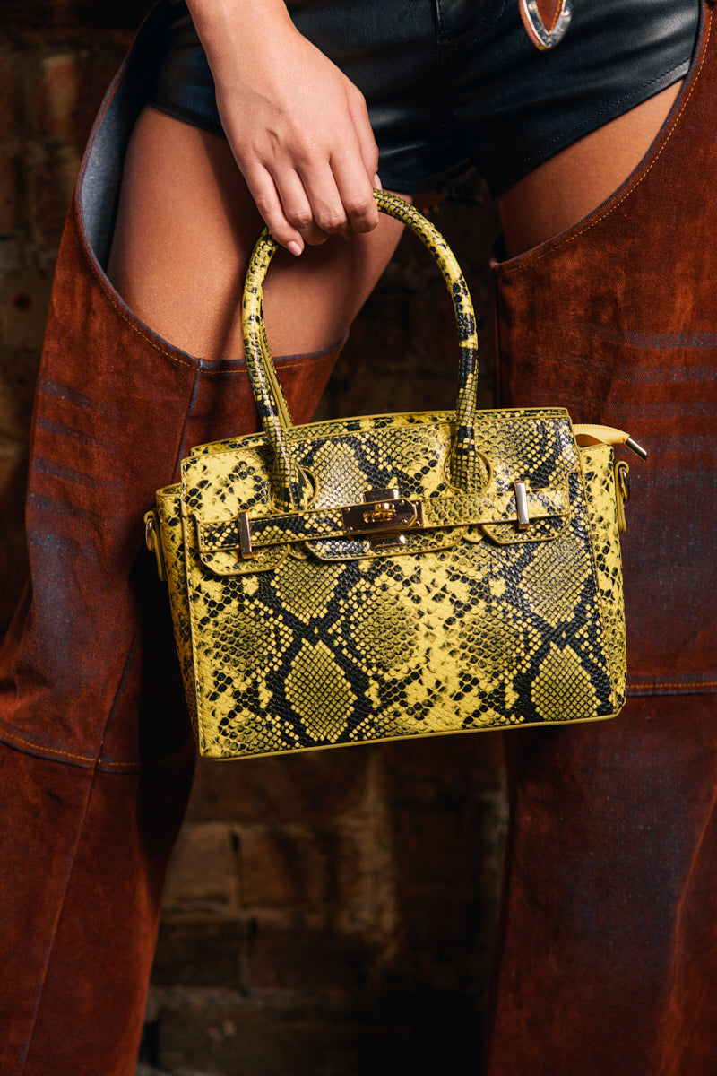 A person in black shorts and brown chaps holds the NEVER TYPICAL SNAKE PRINT PURSE, featuring gold hardware, in front of a rustic wooden background.