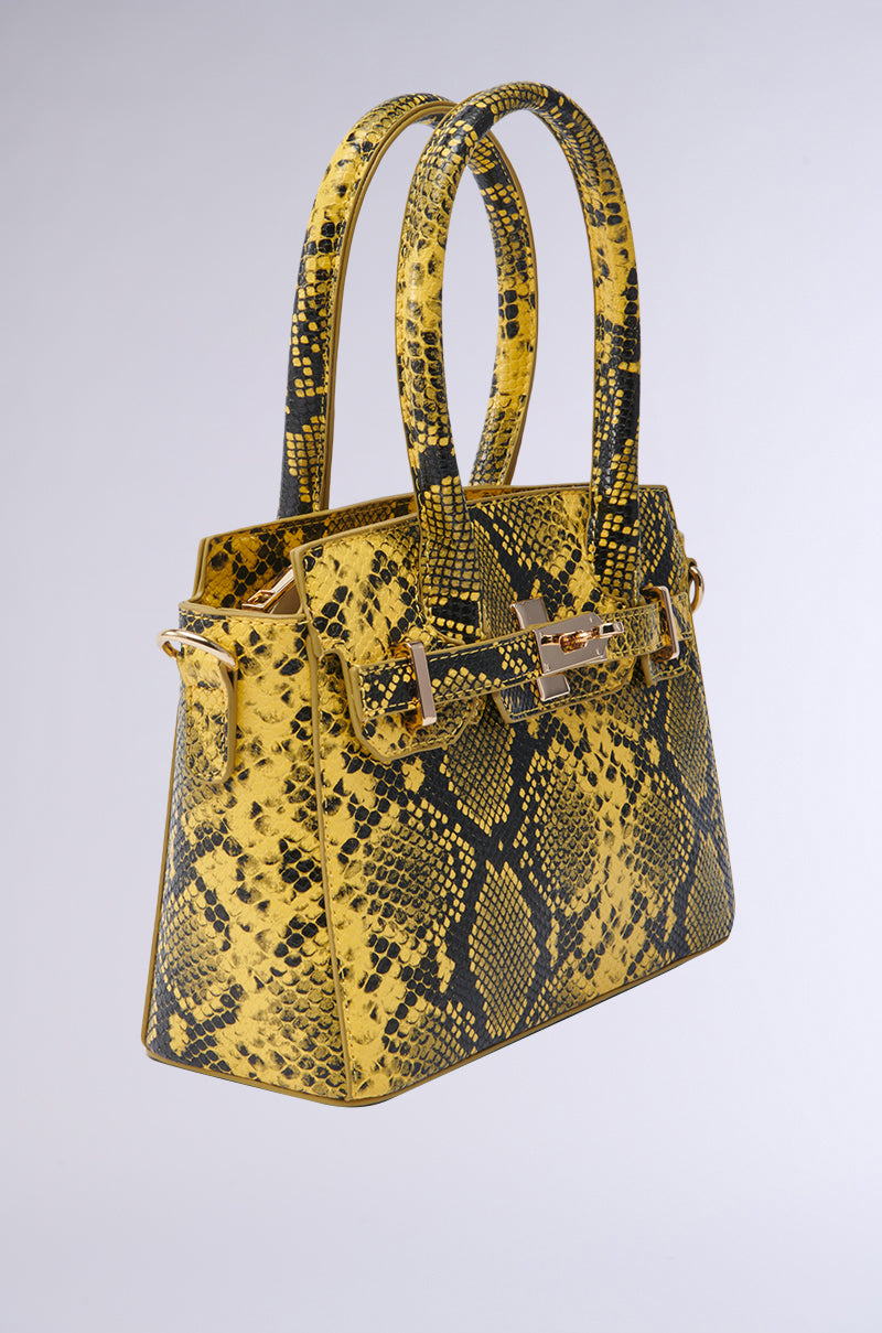 The NEVER TYPICAL SNAKE PRINT PURSE features a yellow and black snakeskin pattern, two handles, and gold hardware, displayed against a plain light gray background.