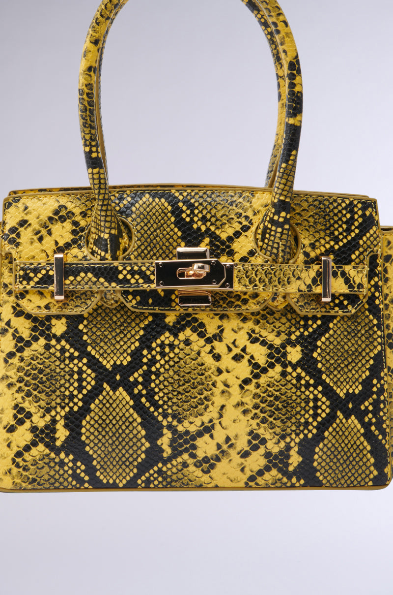 The NEVER TYPICAL SNAKE PRINT PURSE is a yellow handbag with a black snakeskin pattern, gold hardware, double handles, and a front flap with a gold turn-lock closure, displayed against a plain light background.