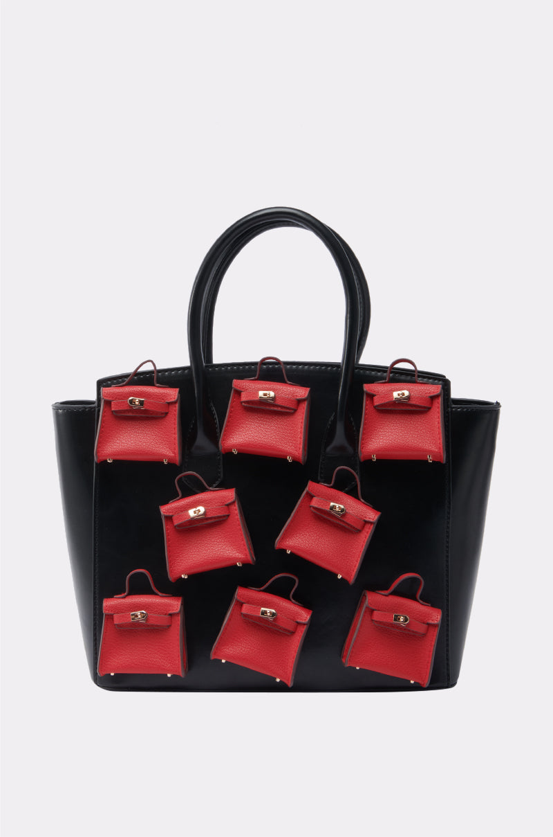 The STAY IN MY BAG PURSE has a structured silhouette in glossy faux leather, with seven small red handbags—each with a handle and gold clasp—arranged in two rows on the front of the large black bag.