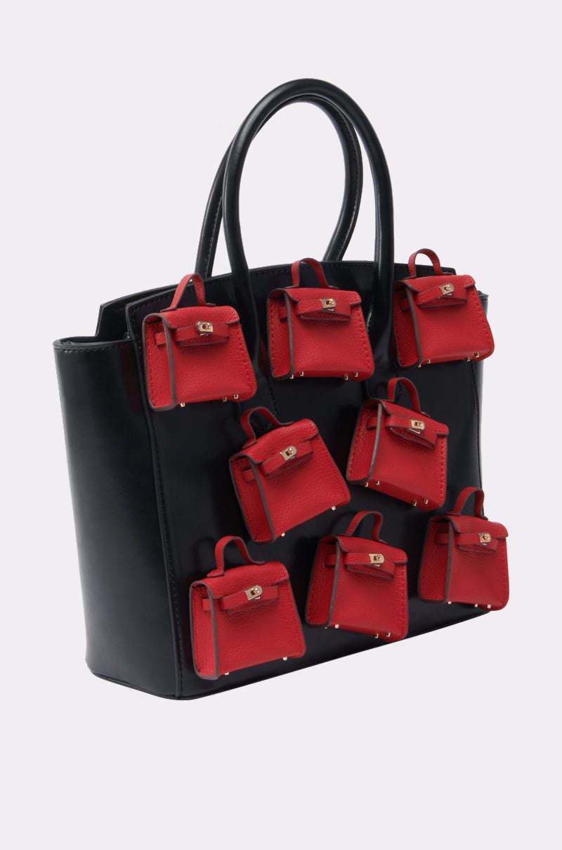 A structured, large black faux leather handbag with a glossy finish, decorated with several small red STAY IN MY BAG PURSE accessories featuring gold clasps and handles, is displayed on a plain light background.