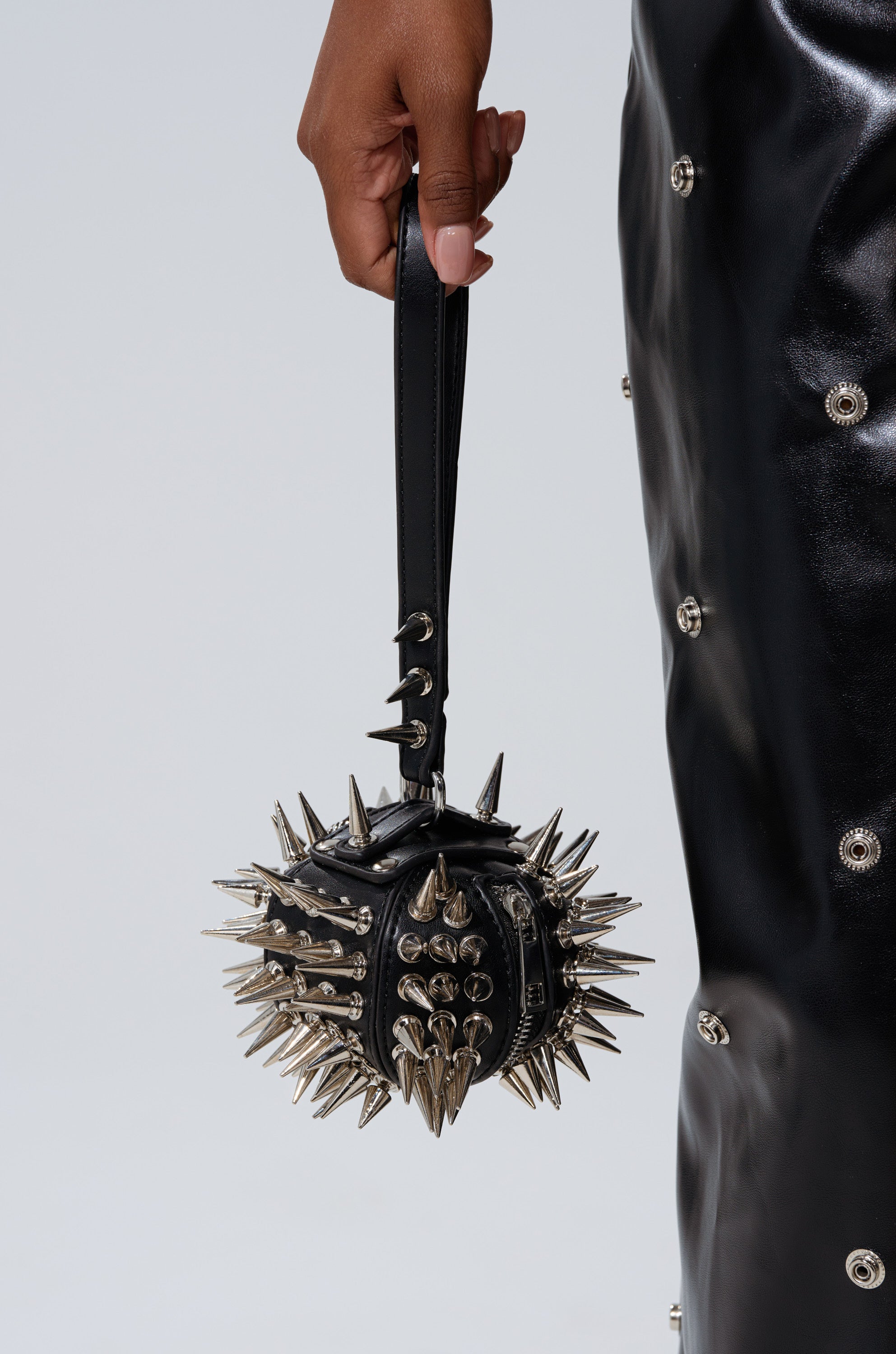 A hand holds the IM SHARP MINI BAG, a silver spike-studded faux leather mini bag with a matching black strap, paired with a shiny black skirt detailed with metal eyelets. The bag stands out against the plain, light-colored background.
