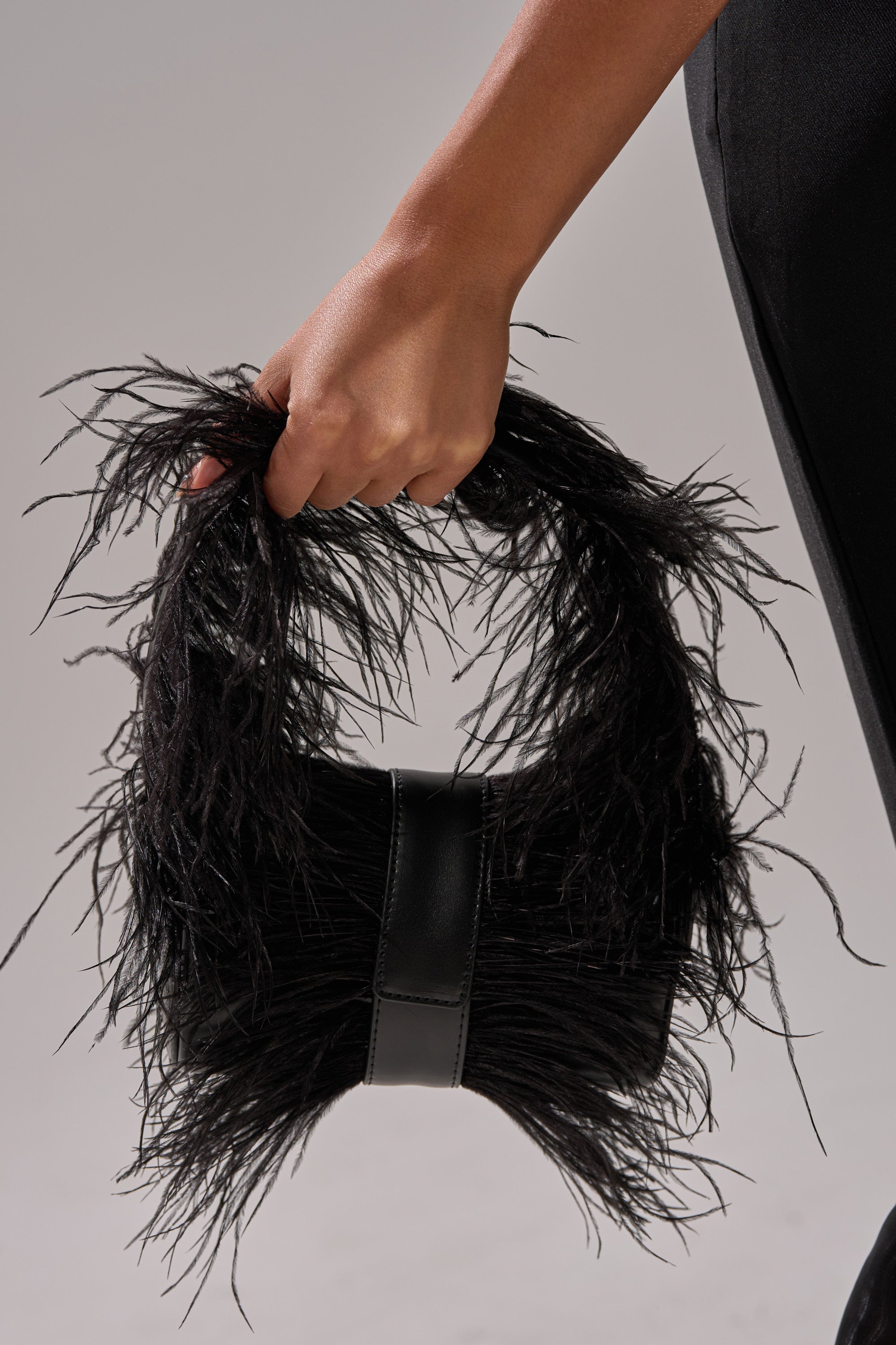 A hand holds the FEATHER CRAZE PURSE—a stylish black feathered bag with bold fringes—against a neutral background. Only the hand, partial arm, and lower black pants show, highlighting this chic evening accessory.