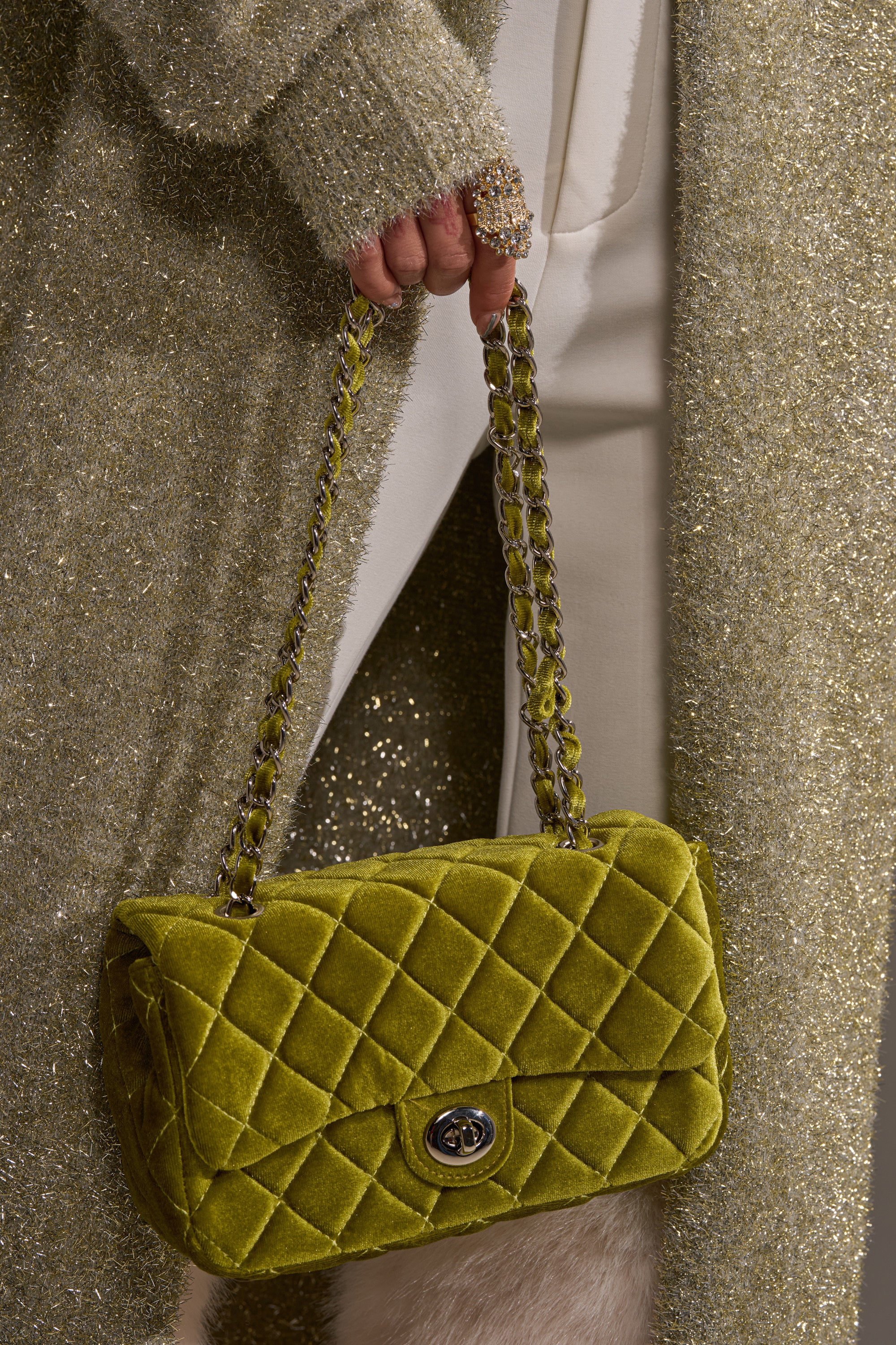 Dressed in glittery gold, a person holds the NIKKI VELVET QUILTED PURSE in olive-green, featuring a chain strap and turn-lock closure. A sparkly gold ring completes the glamorous look.