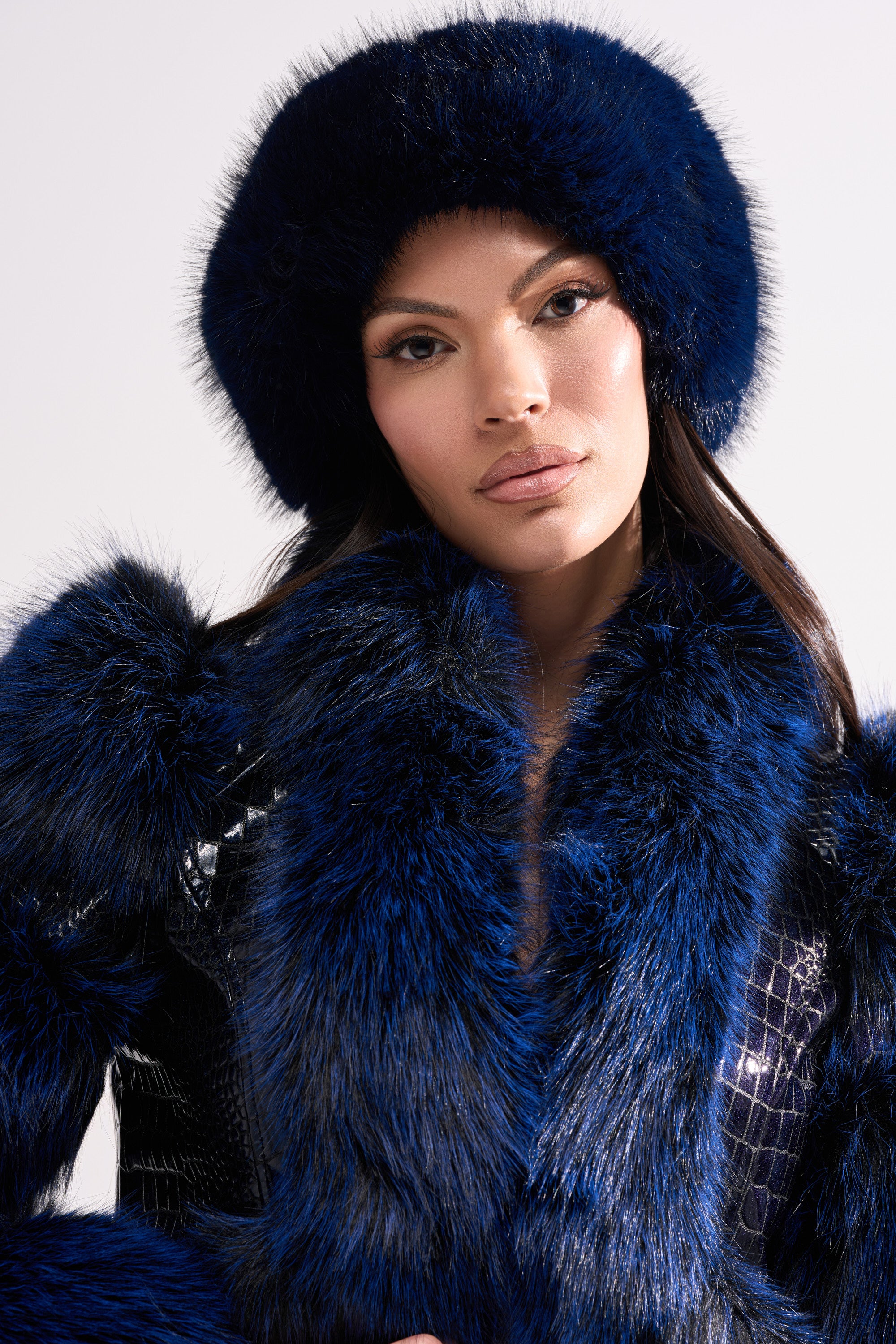 A woman with long dark hair wears the SO CHIC FAUX FUR HEADBAND in deep blue, paired with a matching coat and shiny black textured top, posing against a simple light background with a neutral expression.