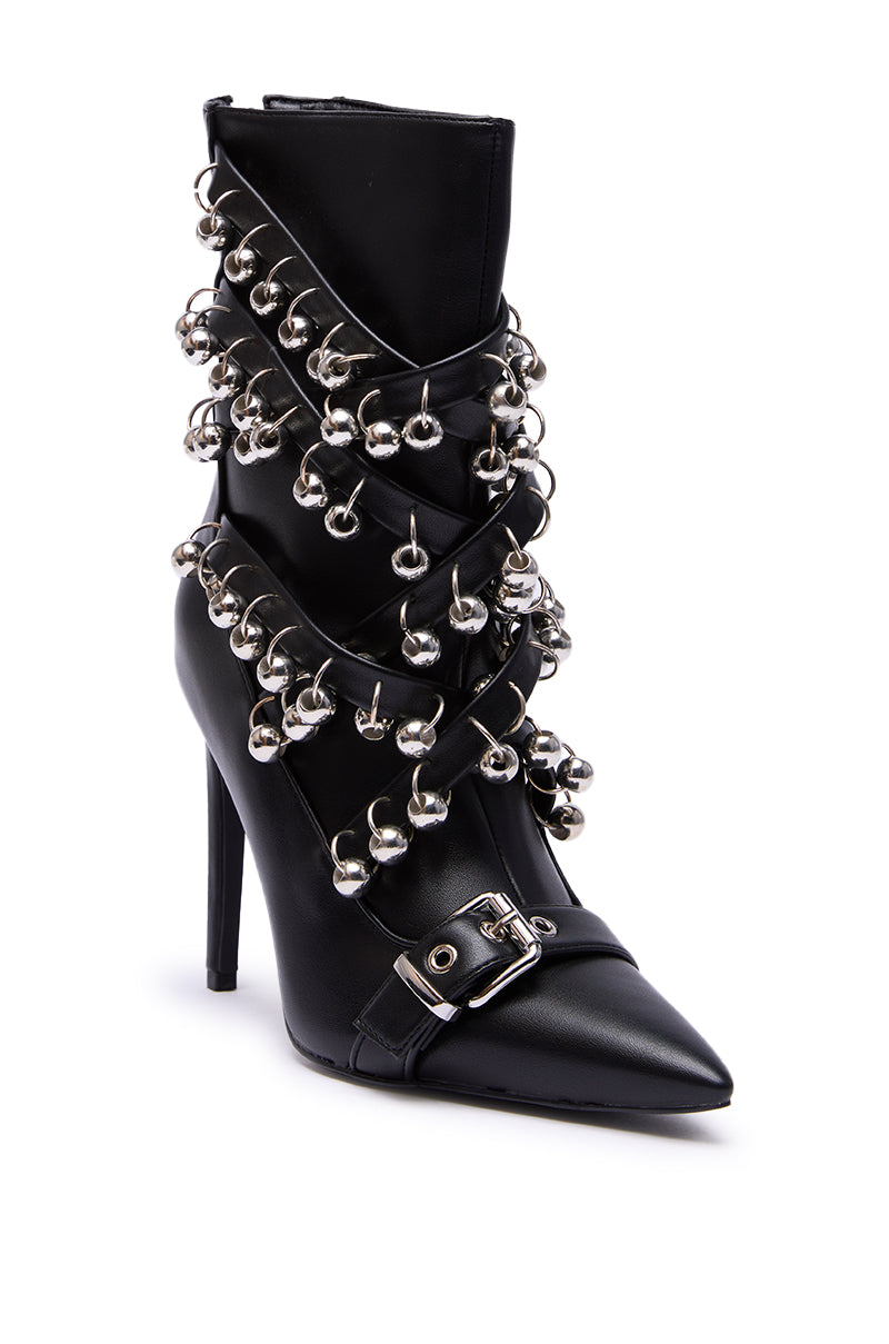 The AZALEA WANG HECATE BLACK METAL boot is a high-heeled black faux leather stiletto featuring a pointed toe, metallic silver jingle bell accents in crisscross patterns, and a chic buckled strap near the toe.