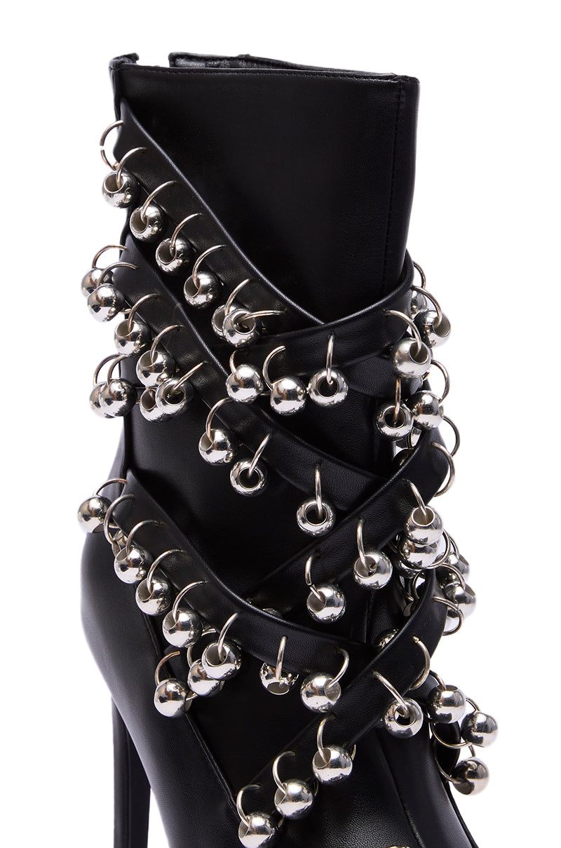 The AZALEA WANG HECATE BLACK METAL is a black stiletto boot with bold crisscross straps and large silver ball studs, featuring a pointed toe for an edgy, statement-making look.