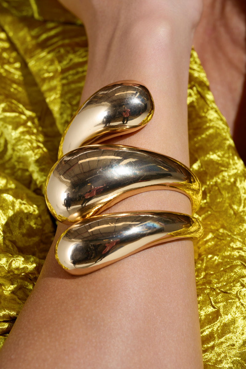 A close-up of the IT IS WHAT IT IS BRACELET CUFF, showcasing smooth, artistic curves spiraling around a forearm. Set against a textured, shiny yellow fabric background, it exudes luxury and elegance.