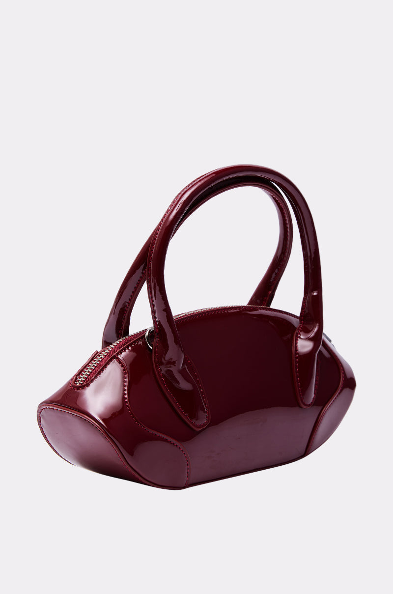 The BELLA PATENT PURSE is a glossy dark red handbag made of patent leather, featuring two rounded handles and a zipper closure, shown against a plain light gray background.