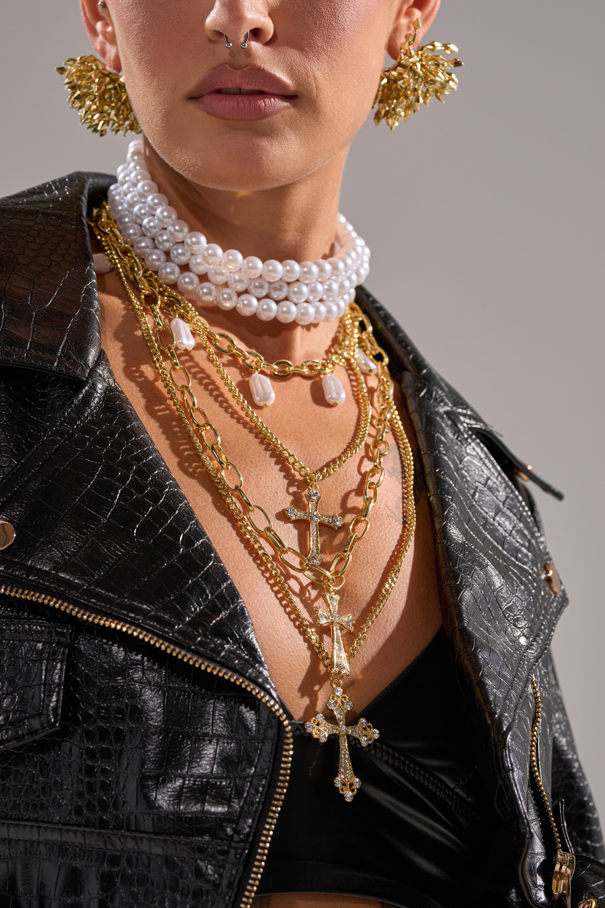 A person wearing a black crocodile-pattern jacket, large gold earrings, a septum piercing, and the TAKE ME ON A TRIP NECKLACE with cross pendants and layered chains. Only their lower face and torso are visible.