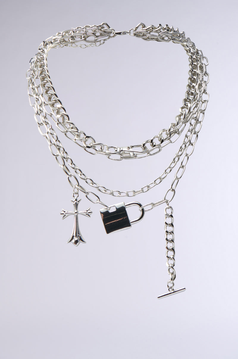 The HEART ON LOCK NECKLACE showcases silver-tone layered chains with a gothic cross pendant and padlock charm in varied styles, beautifully displayed on a light grey background.