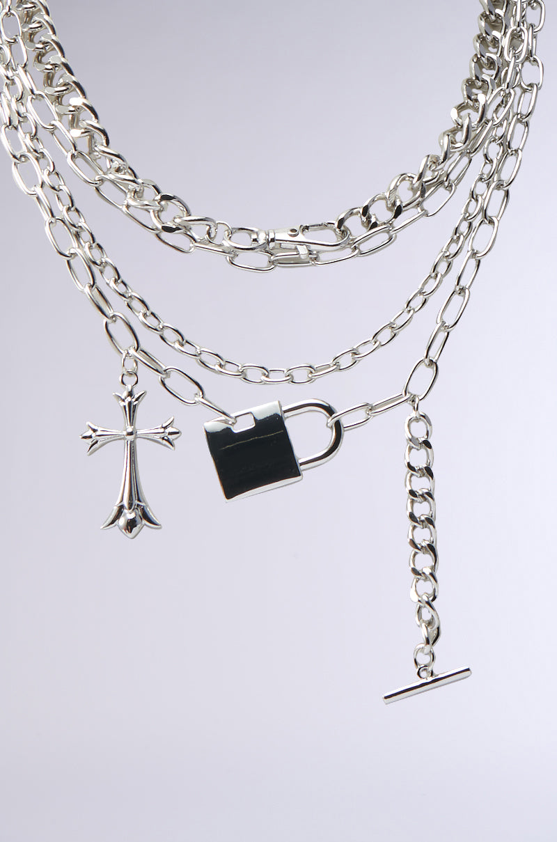 The HEART ON LOCK NECKLACE showcases silver-tone layered chains with a gothic cross pendant, padlock charm, and bar clasp, set against a plain light background.