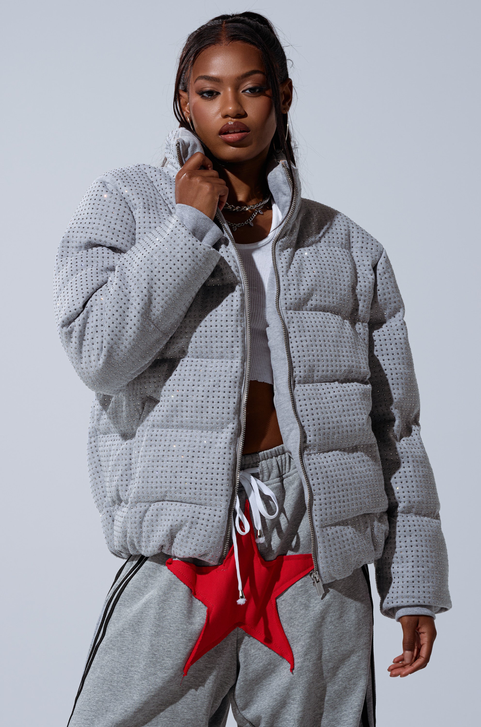 A woman models the HOUDINI MID RHINESTONE PUFFER in light grey over a white crop top and grey sweatpants with red star accents, standing against a plain background.